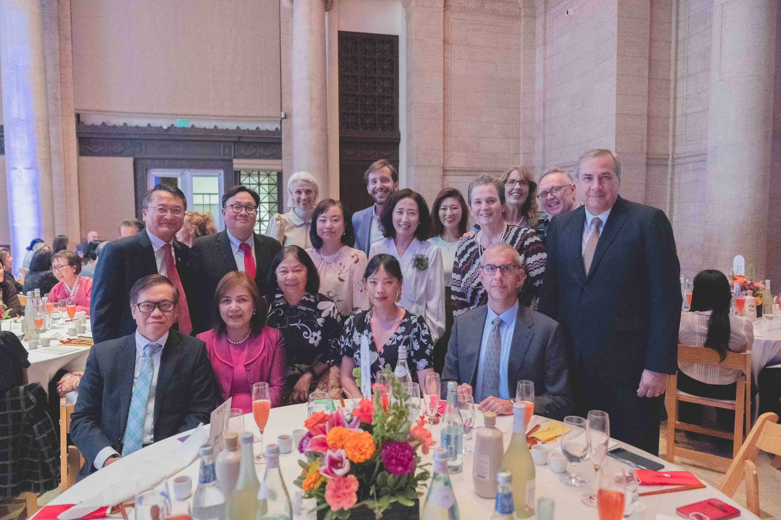 Group of people dressed in formal attire at a banquet or celebration, posing for a photo inside a large hall with high ceilings and stone walls.