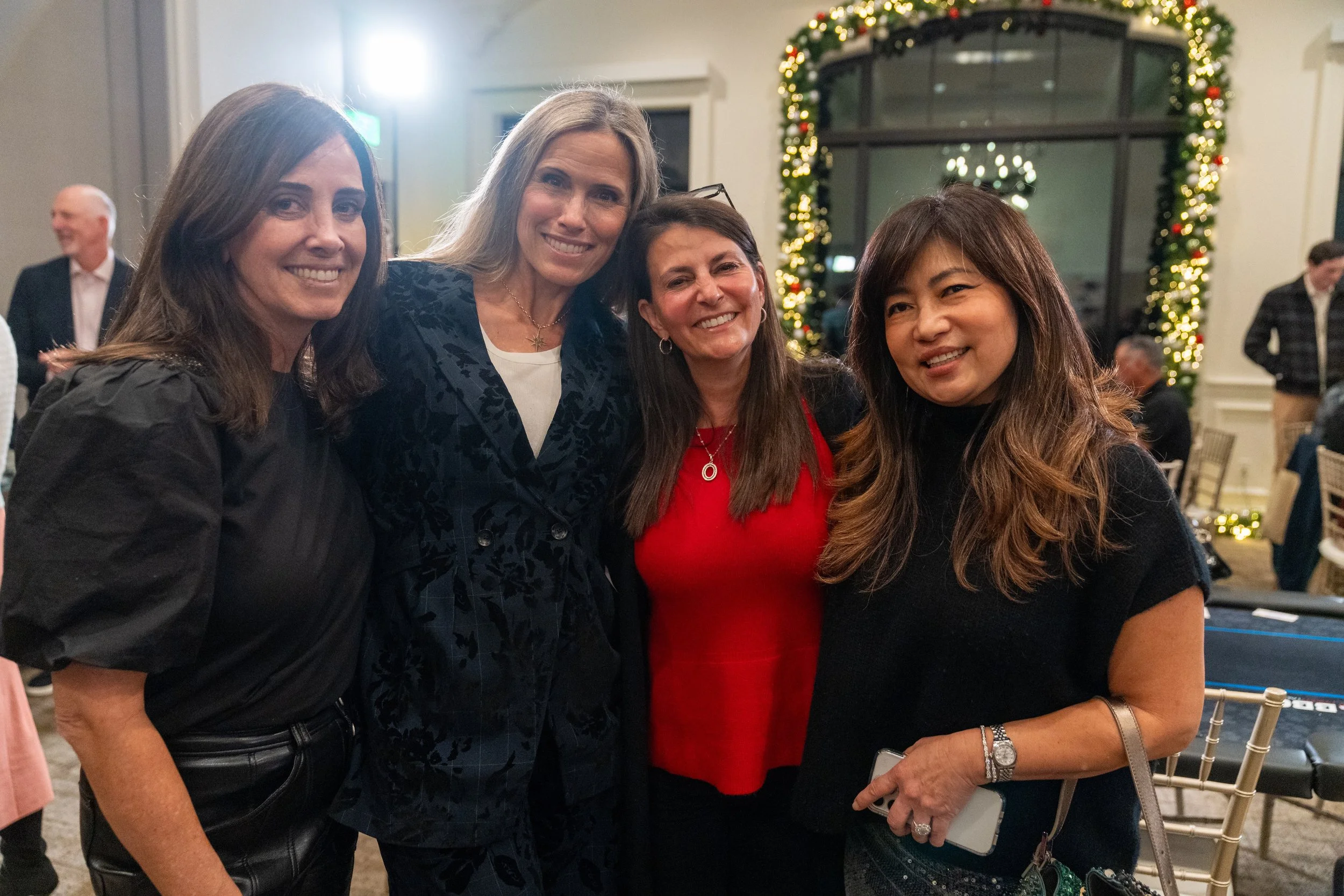 Group of four women smiling and posing at an indoor event with holiday decorations in the background.