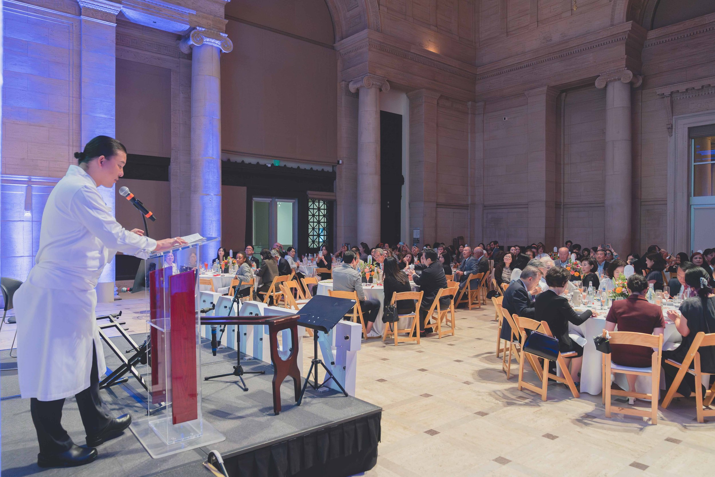A speaker in a white coat stands at a podium on a small stage reading from notes to an audience at round tables in a large, ornate hall.