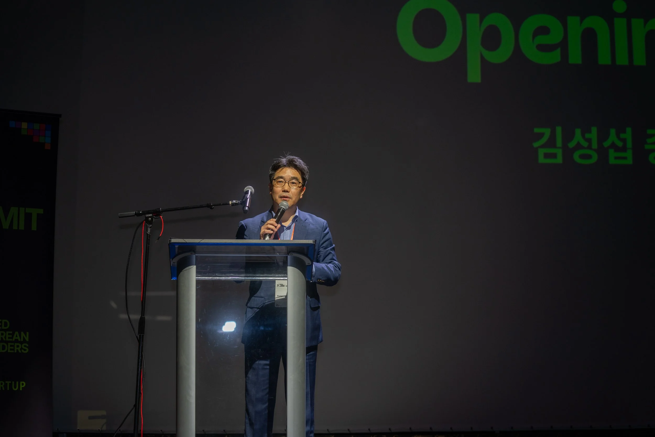 Man speaking at a podium under stage lighting with a microphone, presentation screen and "Opening" text in background.
