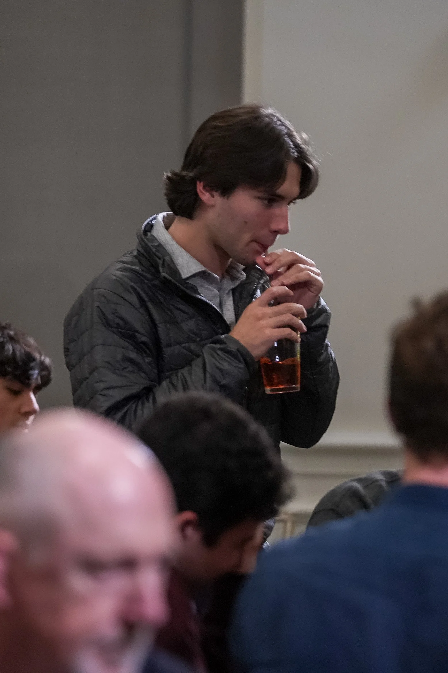 A young man sipping a drink through a straw. He is wearing a dark jacket and standing in a room with other people.