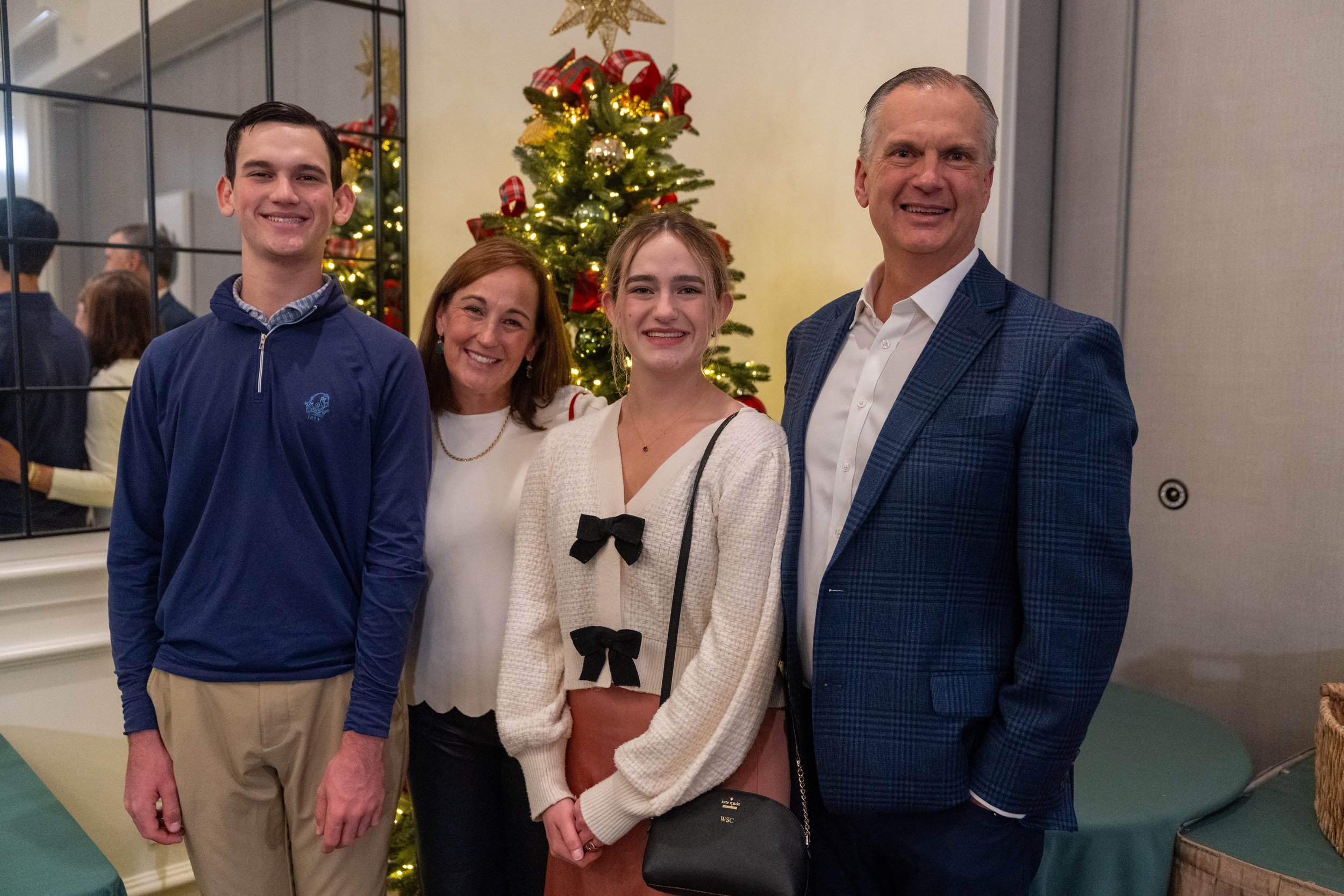 A group of four people standing in front of a decorated Christmas tree, smiling for a photo in a room with a mirror reflecting their image.