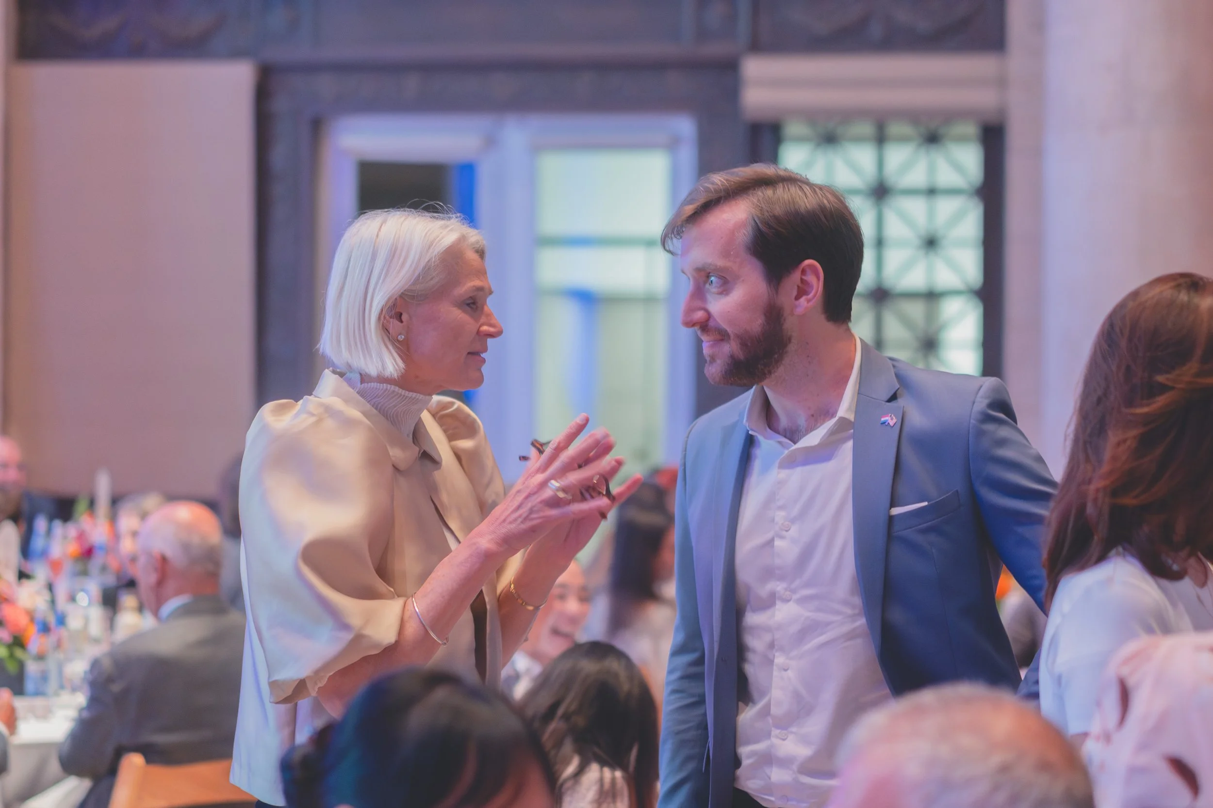 A woman with light hair and a light-colored blouse is talking to a man with dark hair, a beard, and a blue suit at a formal event. They are engaged in conversation among other seated people at tables in a banquet hall.