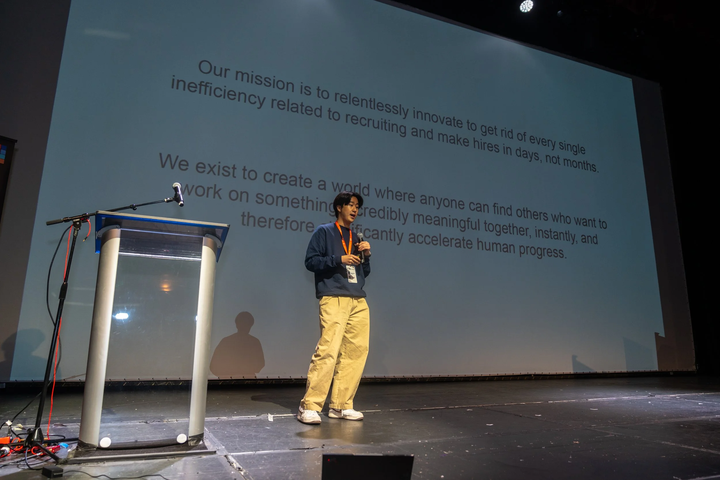 Person presenting on stage in front of a large screen displaying a mission statement. A lectern with a microphone is on the side.