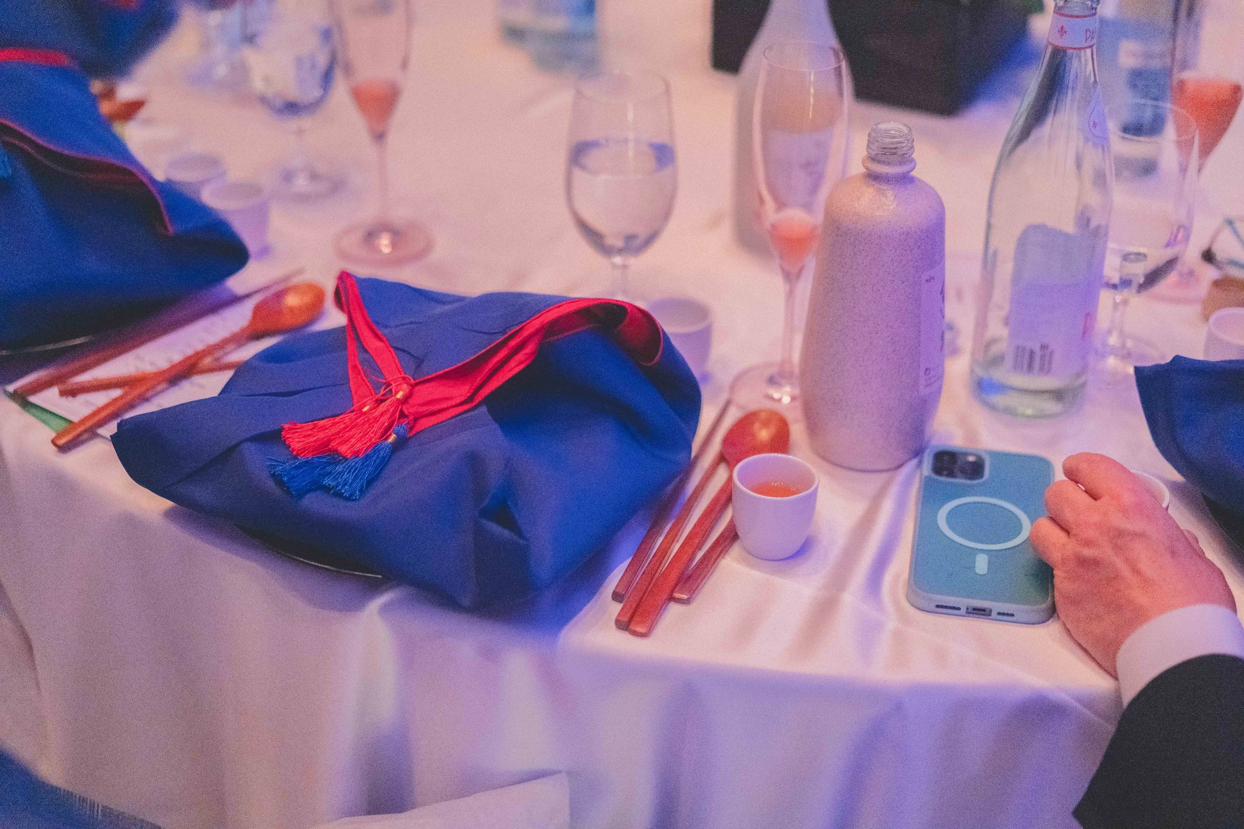 A round banquet table set for a celebration, featuring a blue gift bag with a red and blue tassel, several empty wine glasses, a bottle of water, a small white cup with a drink, a layered ceramic bottle, and a person's hand holding a smartphone.