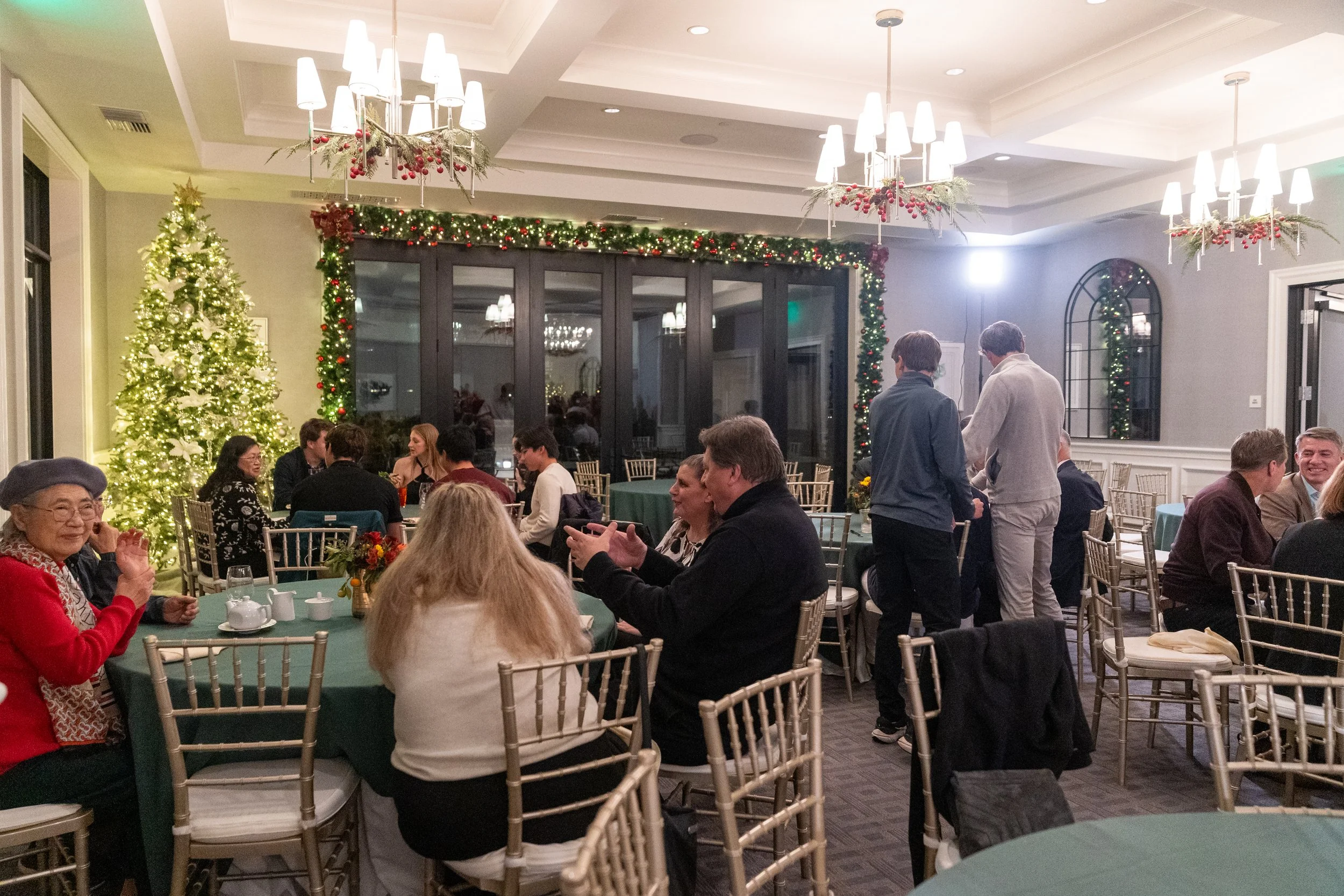 Indoor holiday gathering with people seated at tables, decorated Christmas tree, festive garlands, and chandeliers.