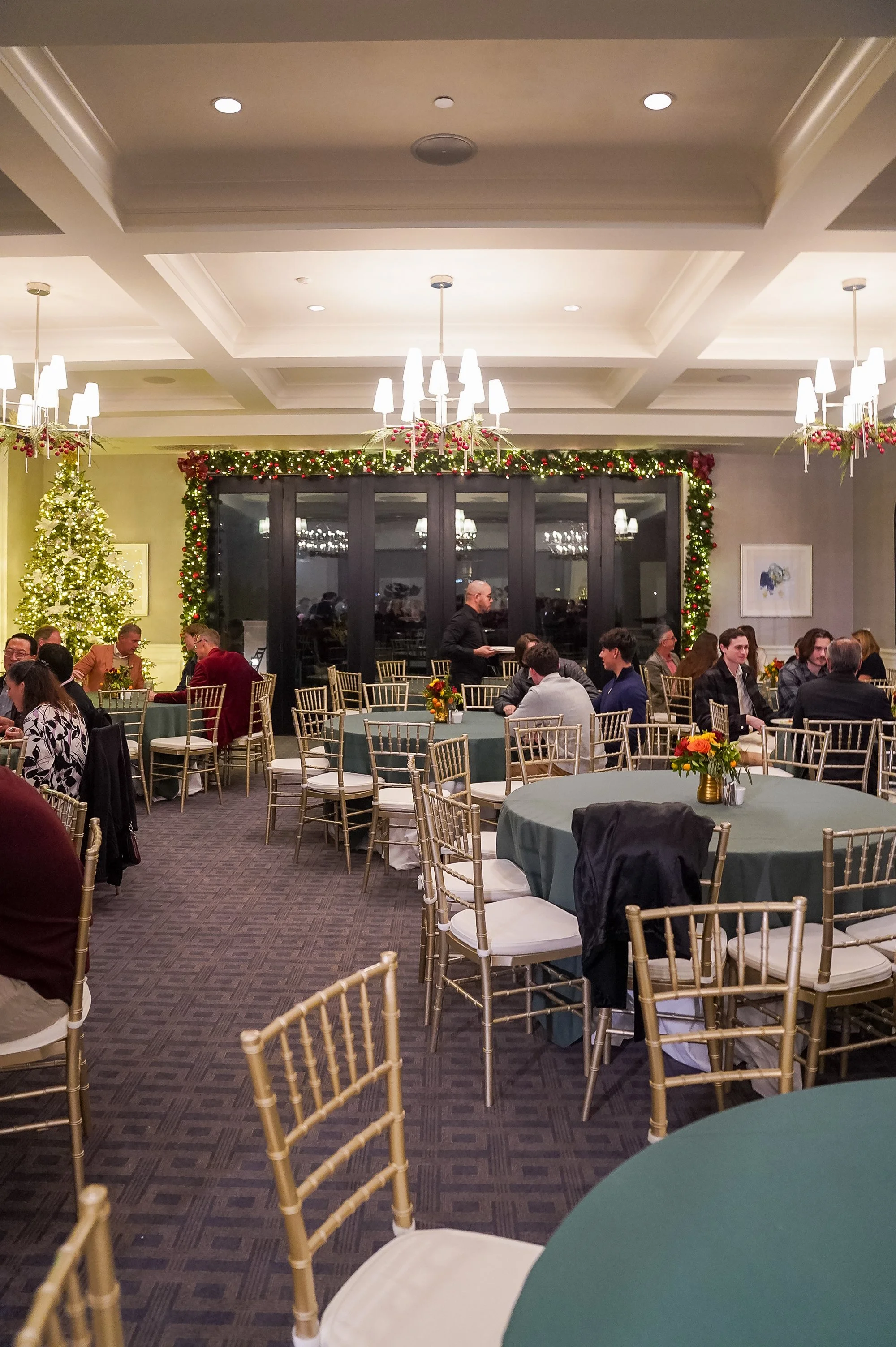 A festive dining hall decorated for the holidays, featuring round tables with green tablecloths and gold chairs. A large Christmas tree and garlands with lights adorn the room. People are seated at the tables, engaged in conversation.