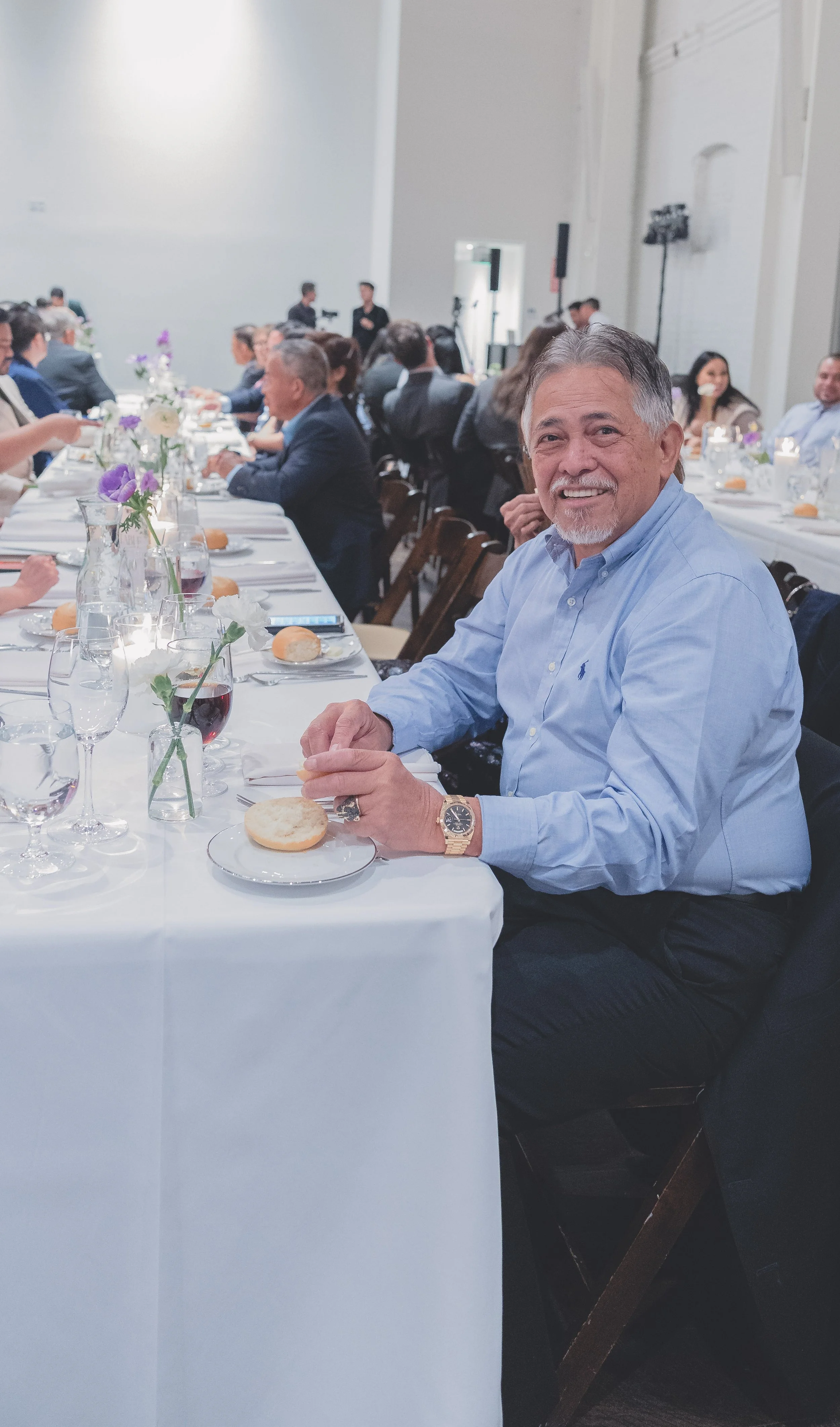 A smiling man in a blue shirt is sitting at a banquet table set with white tablecloth, bread rolls, wine glasses, and floral centerpieces. The dining room is filled with people seated at tables in the background.