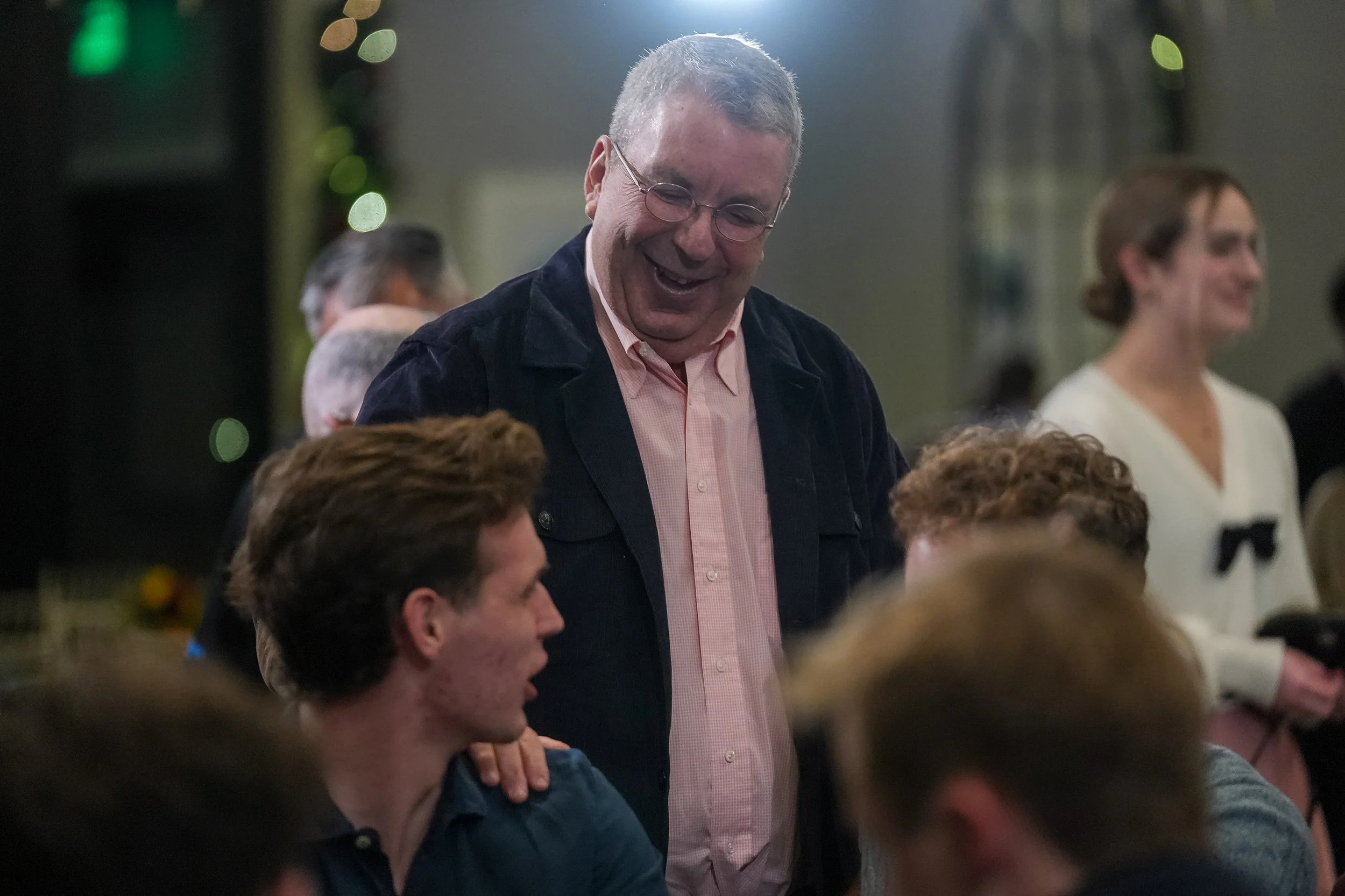 A group of people interacting at a social event indoors. A man wearing glasses and a jacket is smiling and engaging with seated individuals, while others chat in the background.