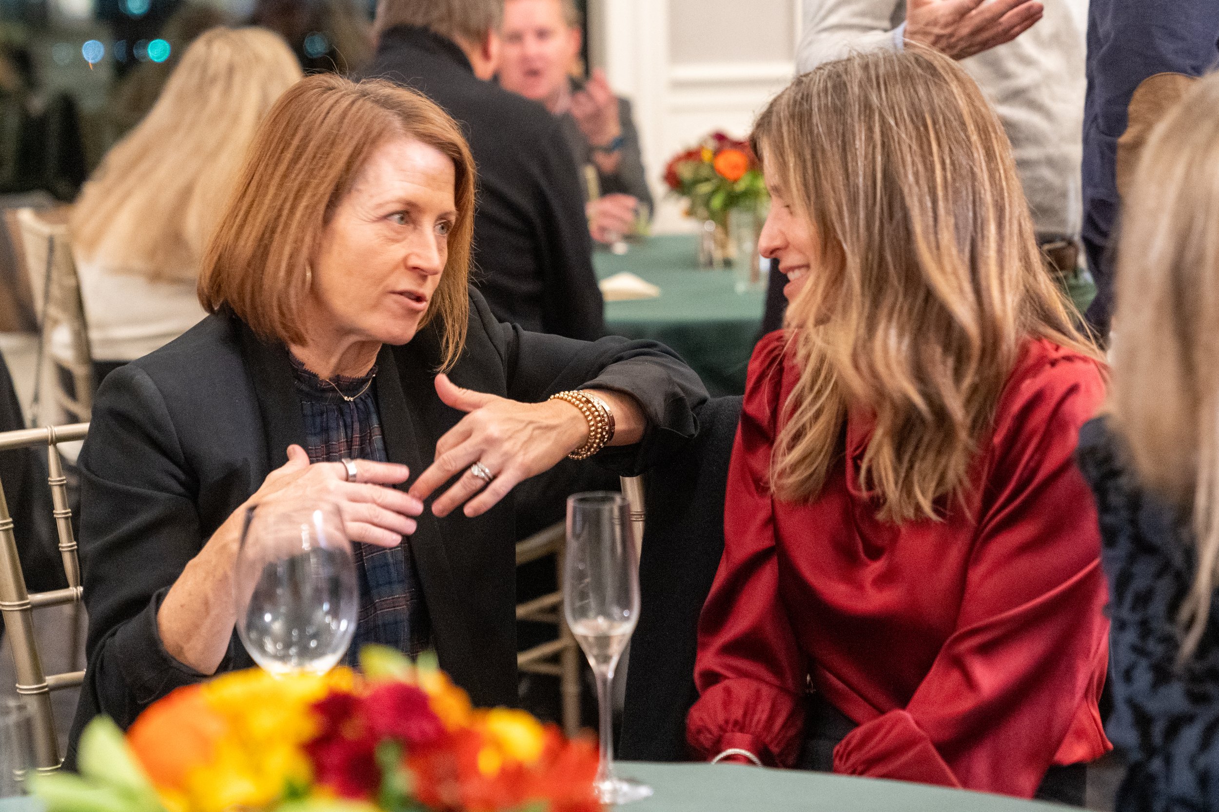 Two women seated at a table, engaged in conversation, with empty wine glasses and floral decorations around them at a social event.