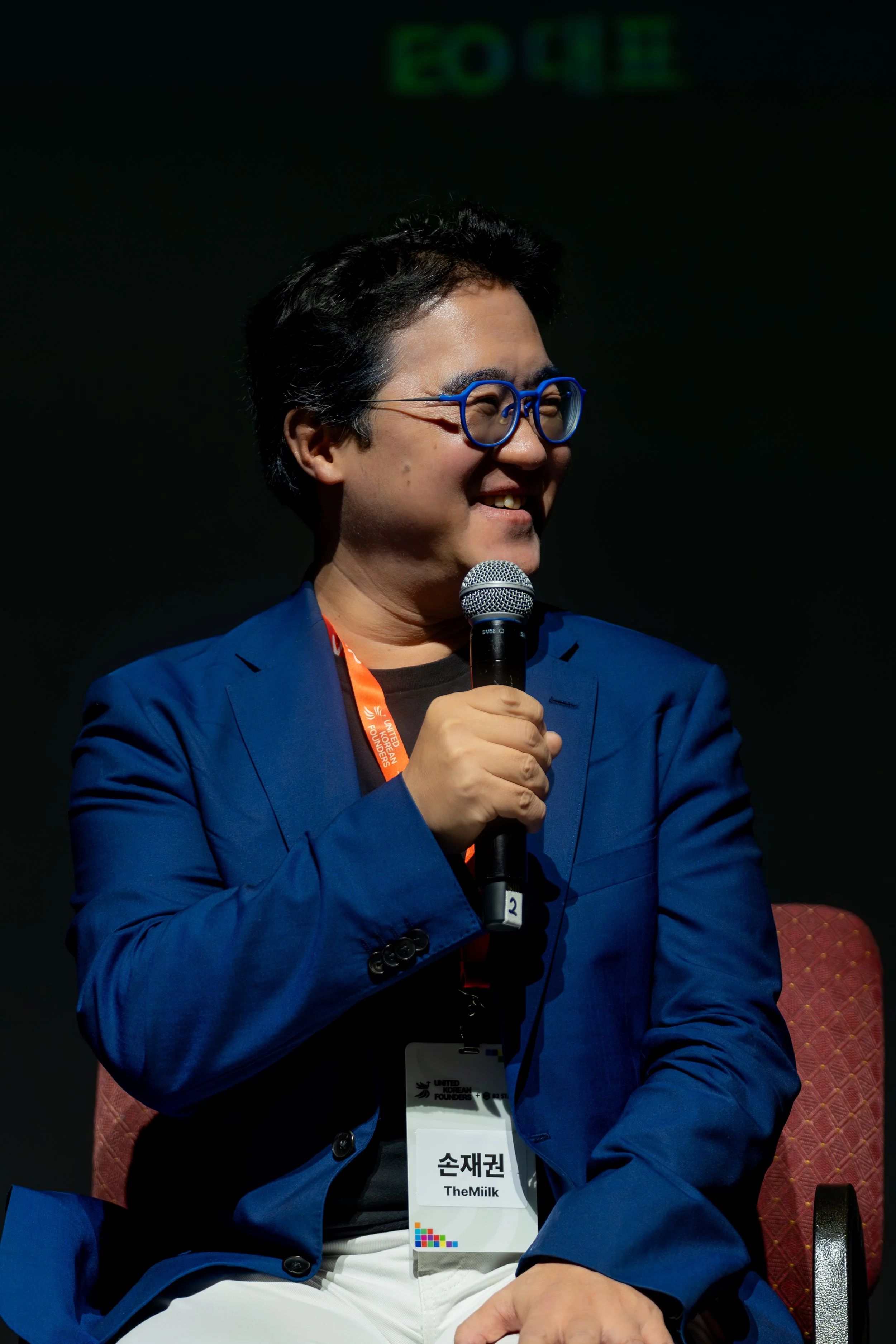 Person in a blue blazer holding a microphone, wearing glasses and a name badge, sitting on a chair.