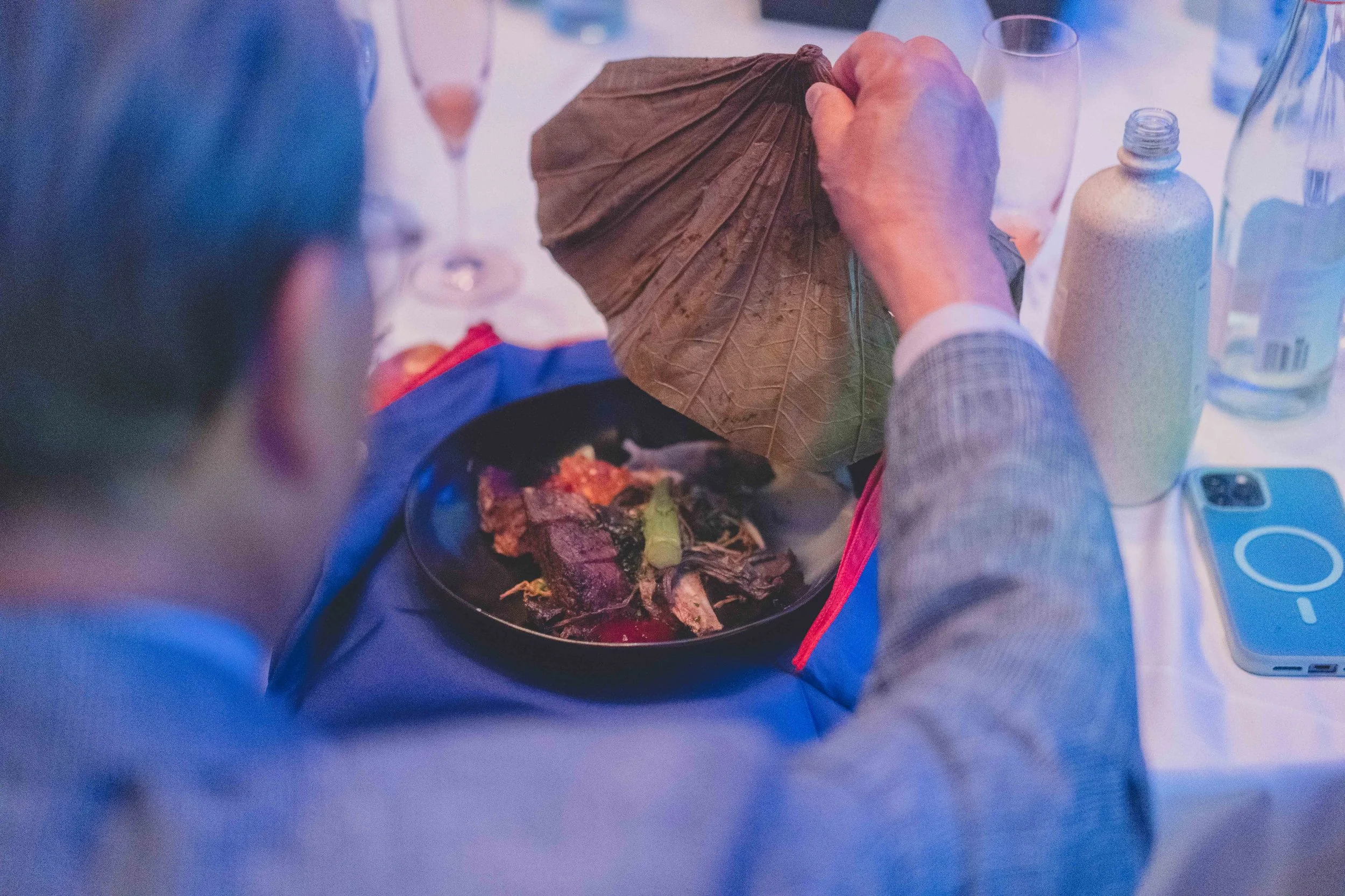 Person holding a dry leaf over a bowl of food on a table with drinks and a smartphone.