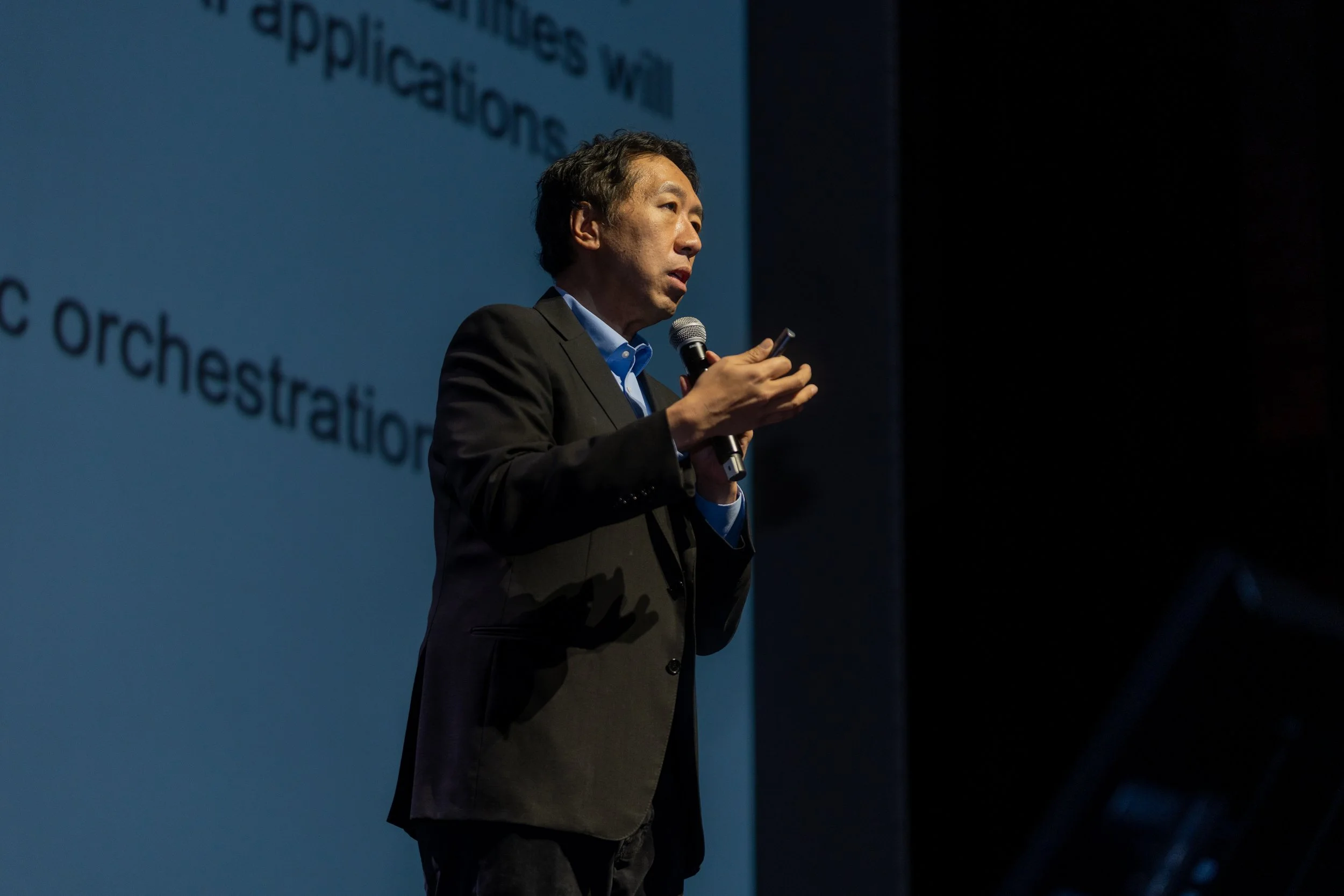 Person speaking on stage holding a microphone with text projected in the background.
