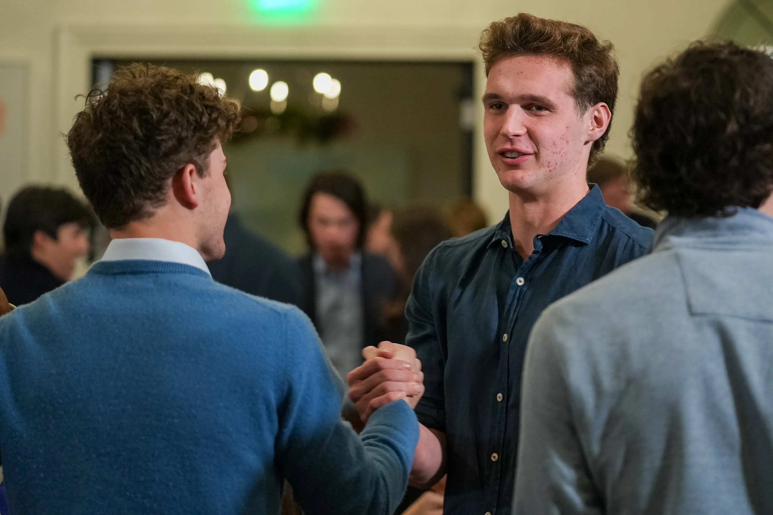 Two people shaking hands at an event, surrounded by a group in a social setting.