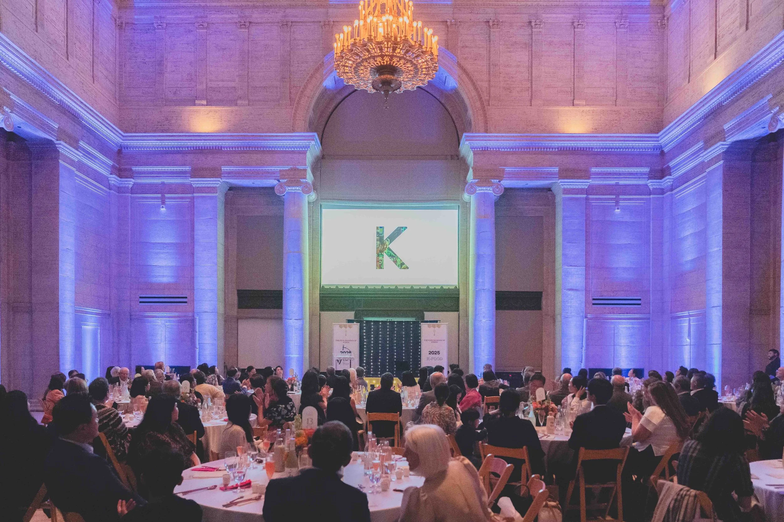 Large banquet hall with high ceilings, purple and warm lighting, a grand chandelier, and a crowd of people seated at round tables, watching a presentation on a large screen with a letter 'K' and decorated banners.
