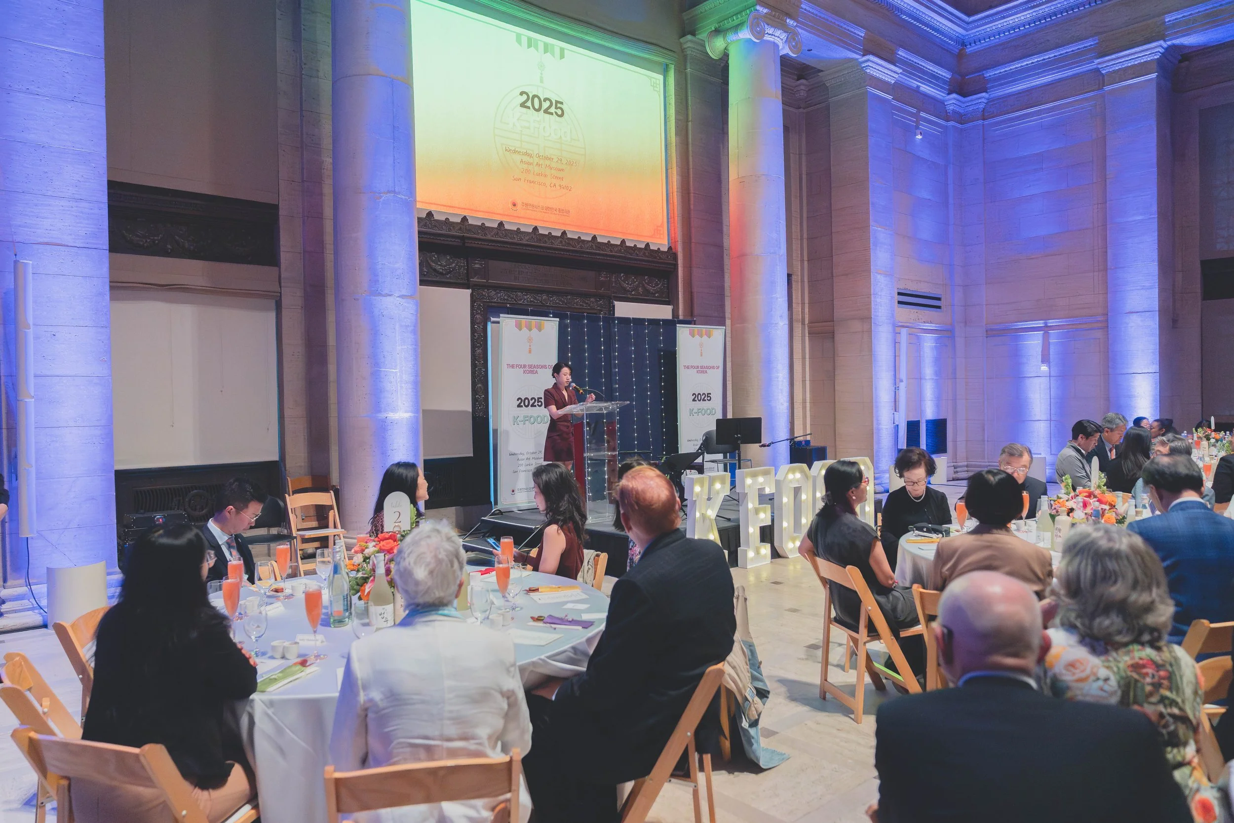 People attending a formal event or conference in a large hall, seated at round tables with floral centerpieces, listening to a speaker on stage with large 