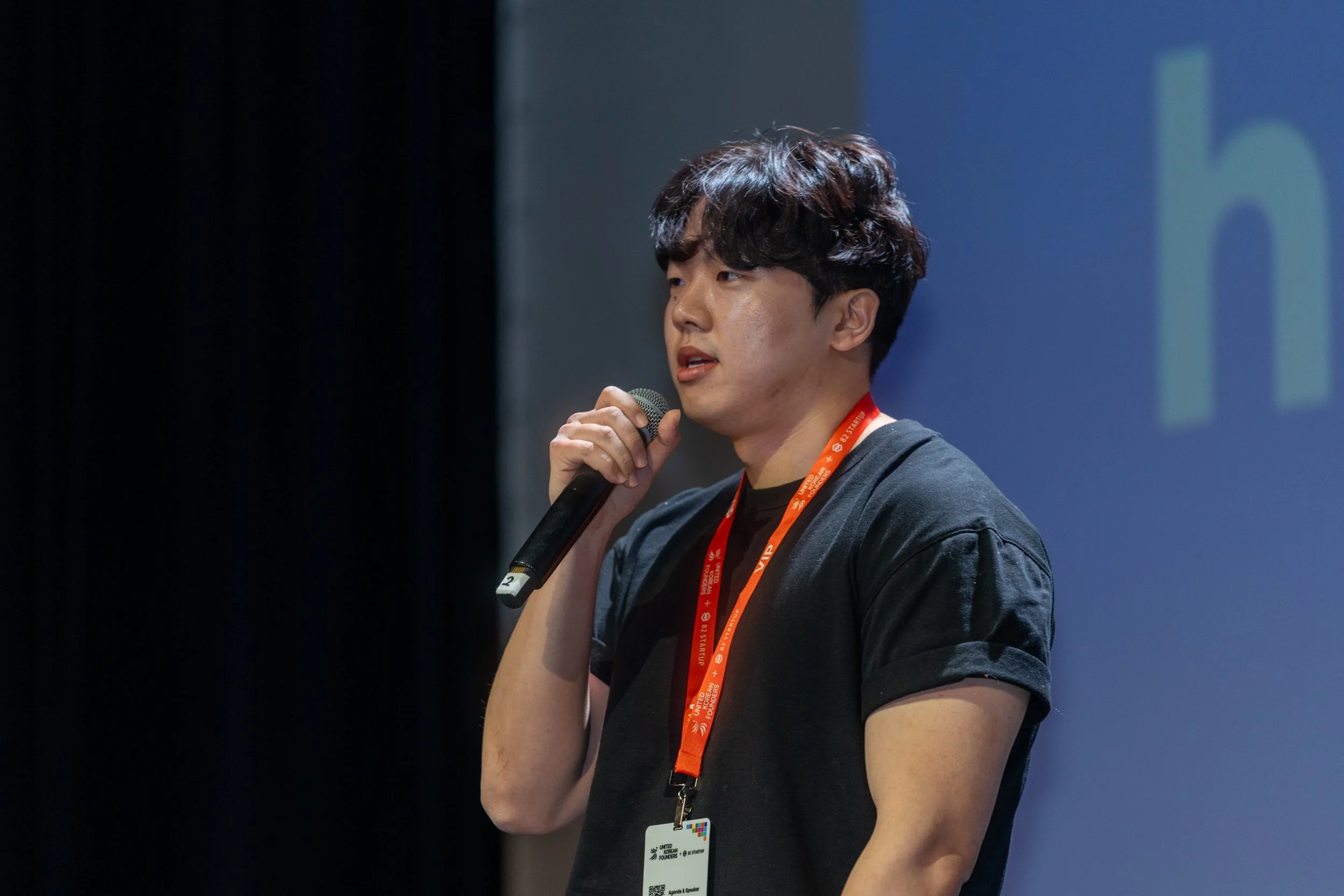 Person speaking into a microphone on stage with a conference badge and lanyard