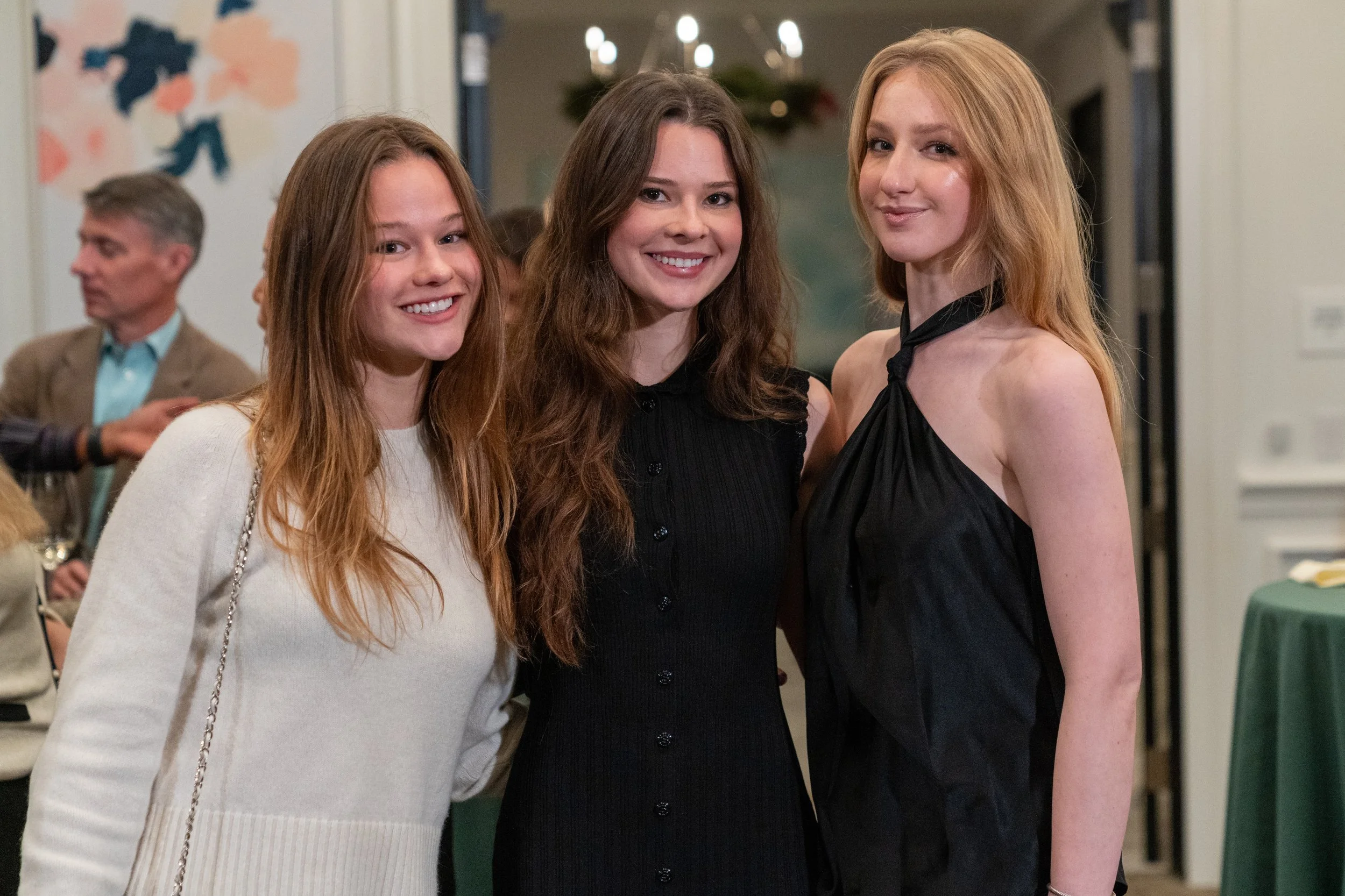 Three young women smiling at a formal event, with people and decorative elements in the background.
