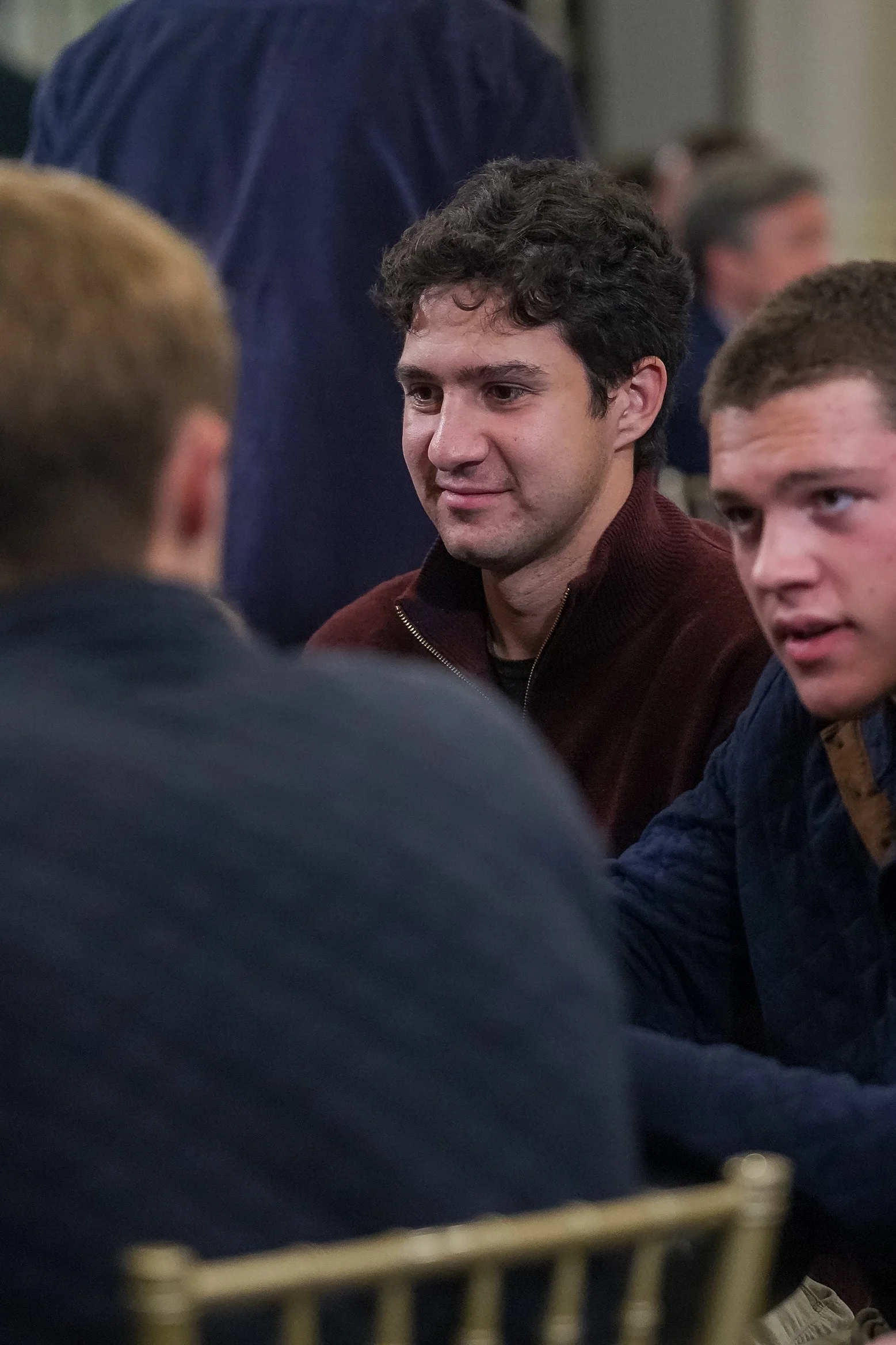 Three men sitting at a table, engaged in conversation. The focus is on the man in the center with curly hair and a maroon sweater. The environment appears to be a social or casual setting.
