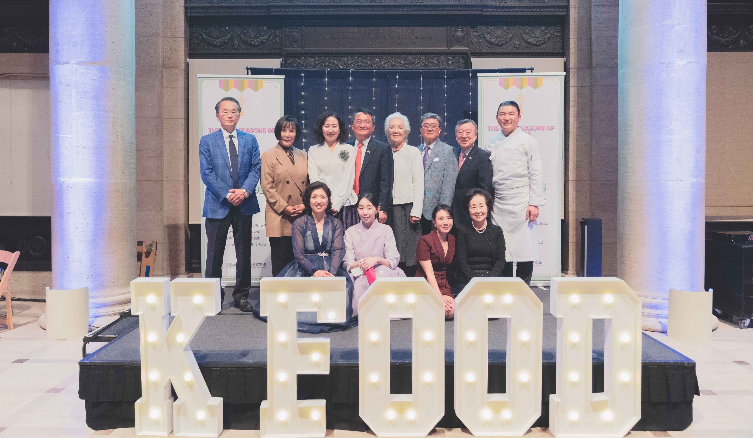 A group of 12 people, some standing and some sitting, posing on a stage behind large illuminated 'KEEP' letters. The background features banners with the text 'THE SEASONS OF' and decorative lighting.