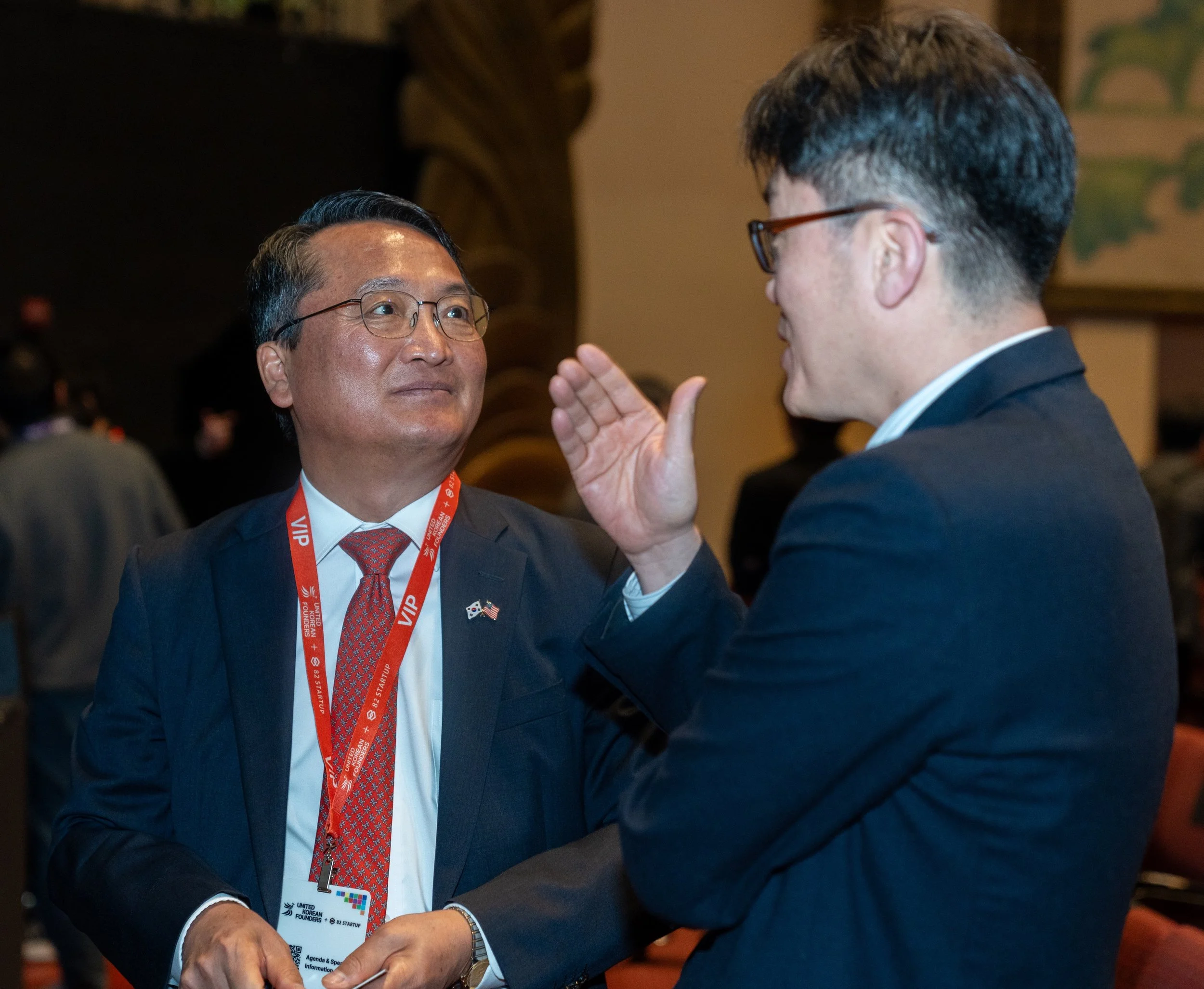Two men in suits with red lanyards having a conversation at an event.