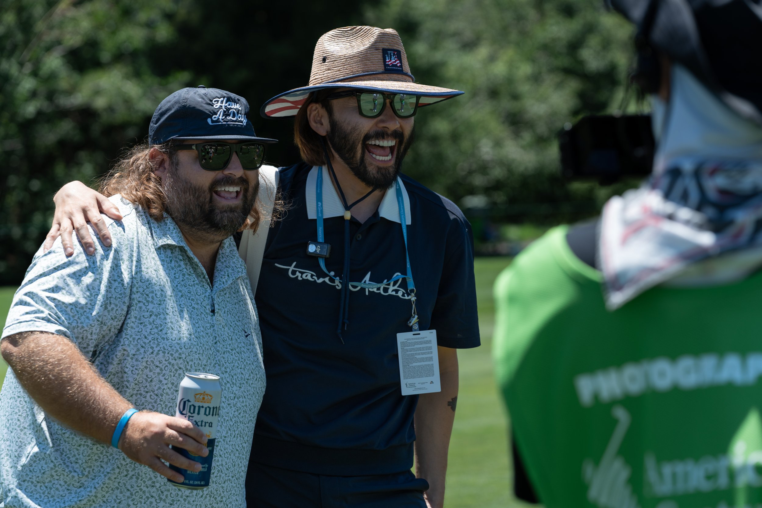 Two men smiling outdoors, one holding a beer can, both wearing sunglasses and casual hats. A photographer in a green vest is in the foreground.