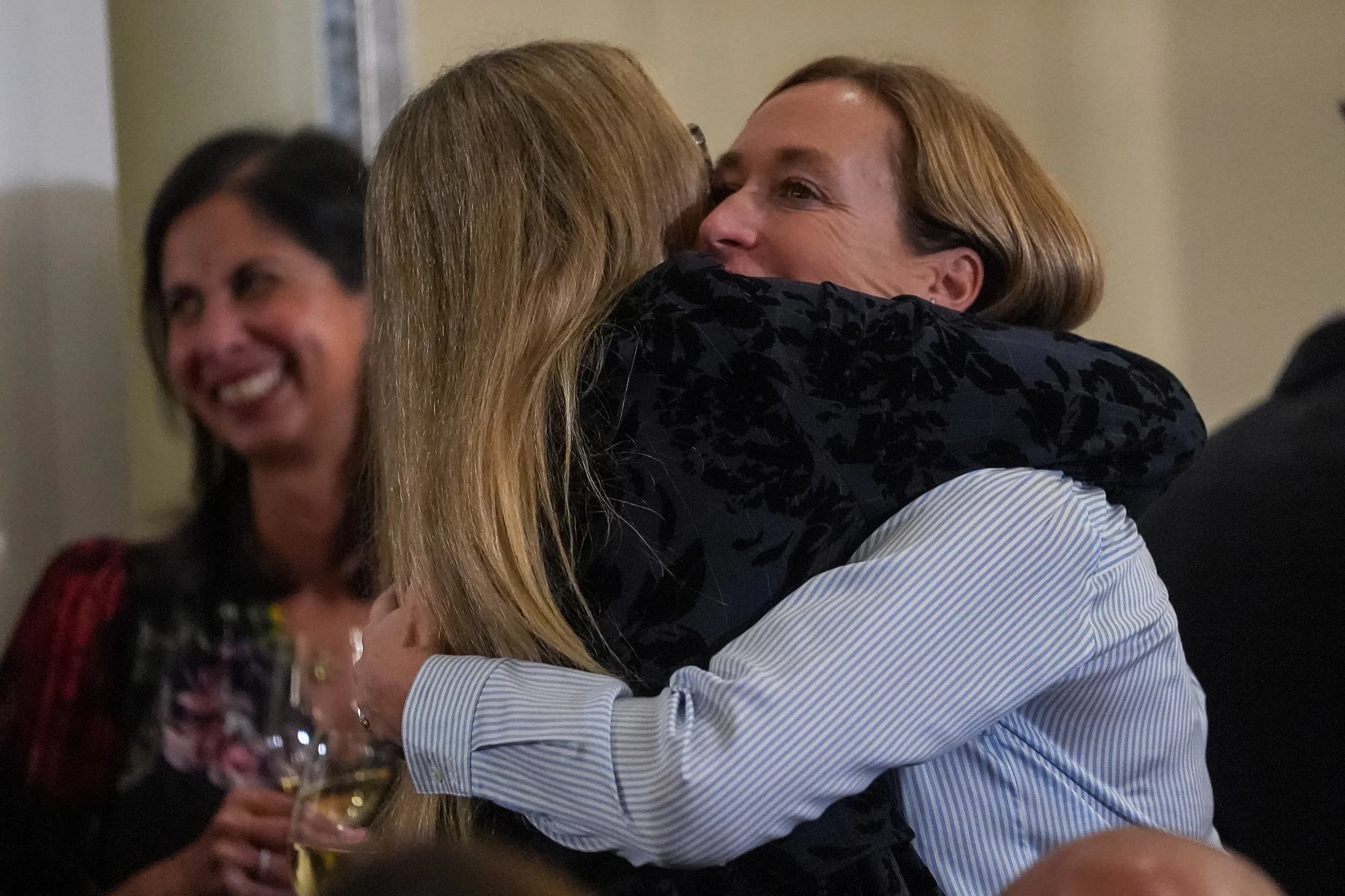 Two women hugging at an event, another woman smiling in the background, holding a glass of wine.