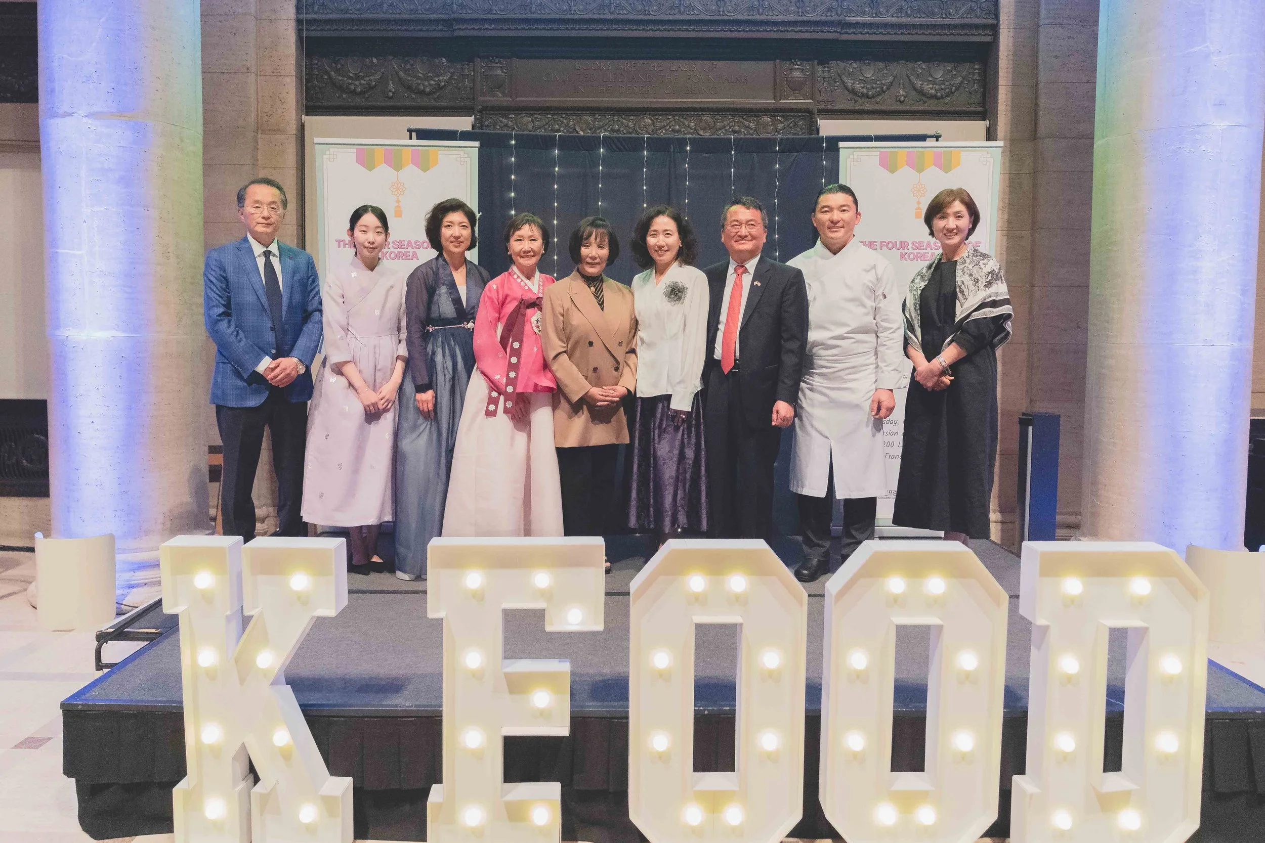 Group of ten people standing on a stage, smiling, with large illuminated letters spelling 'K-FOOD' in front. The background features banners with the text 'The Four Seasons Korea.' The individuals are dressed in formal and traditional Korean attire, 