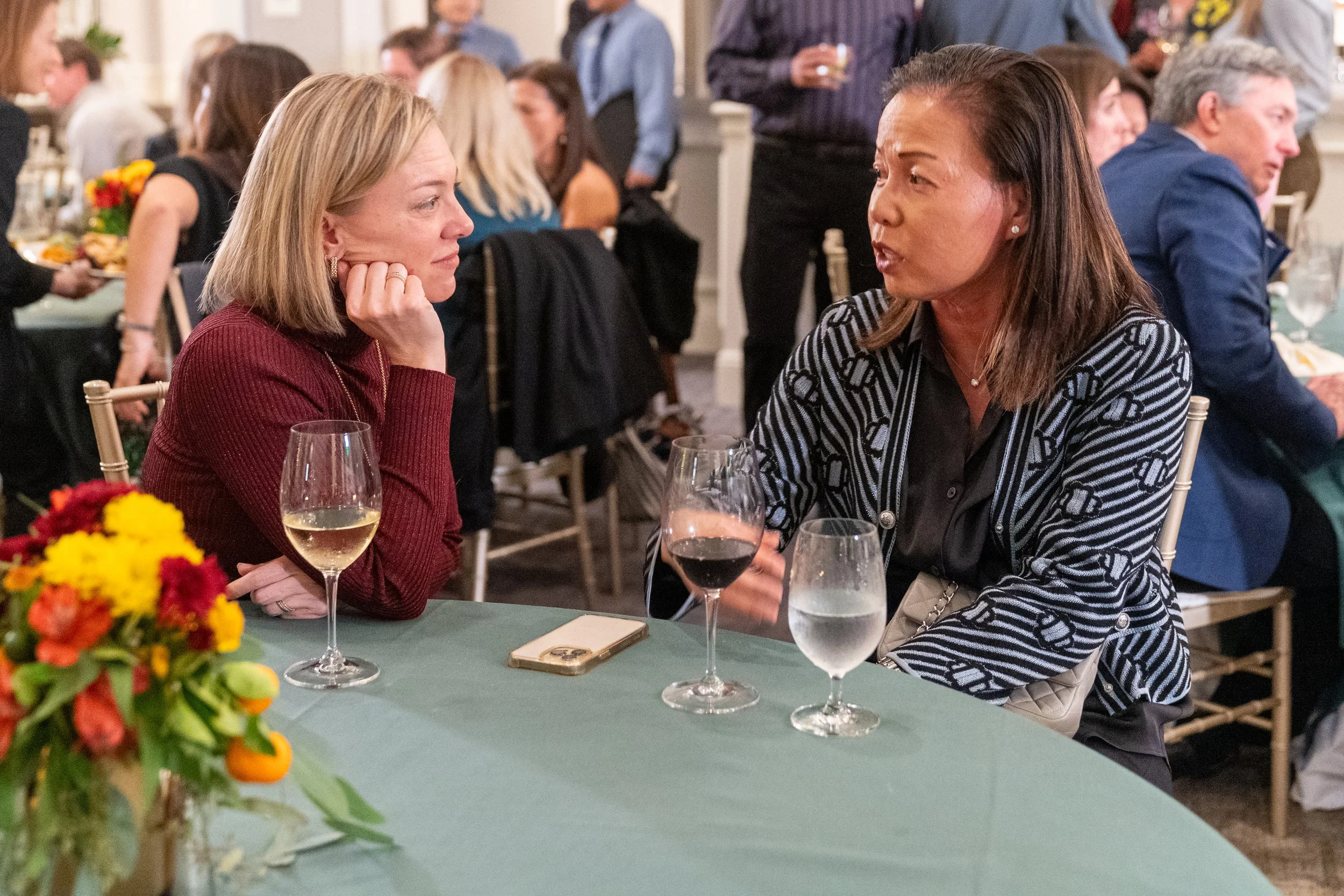 Two women sitting at a table with drinks, engaged in conversation at a social event. The table is decorated with flowers and a smartphone is lying on it. A group of people is visible in the background.
