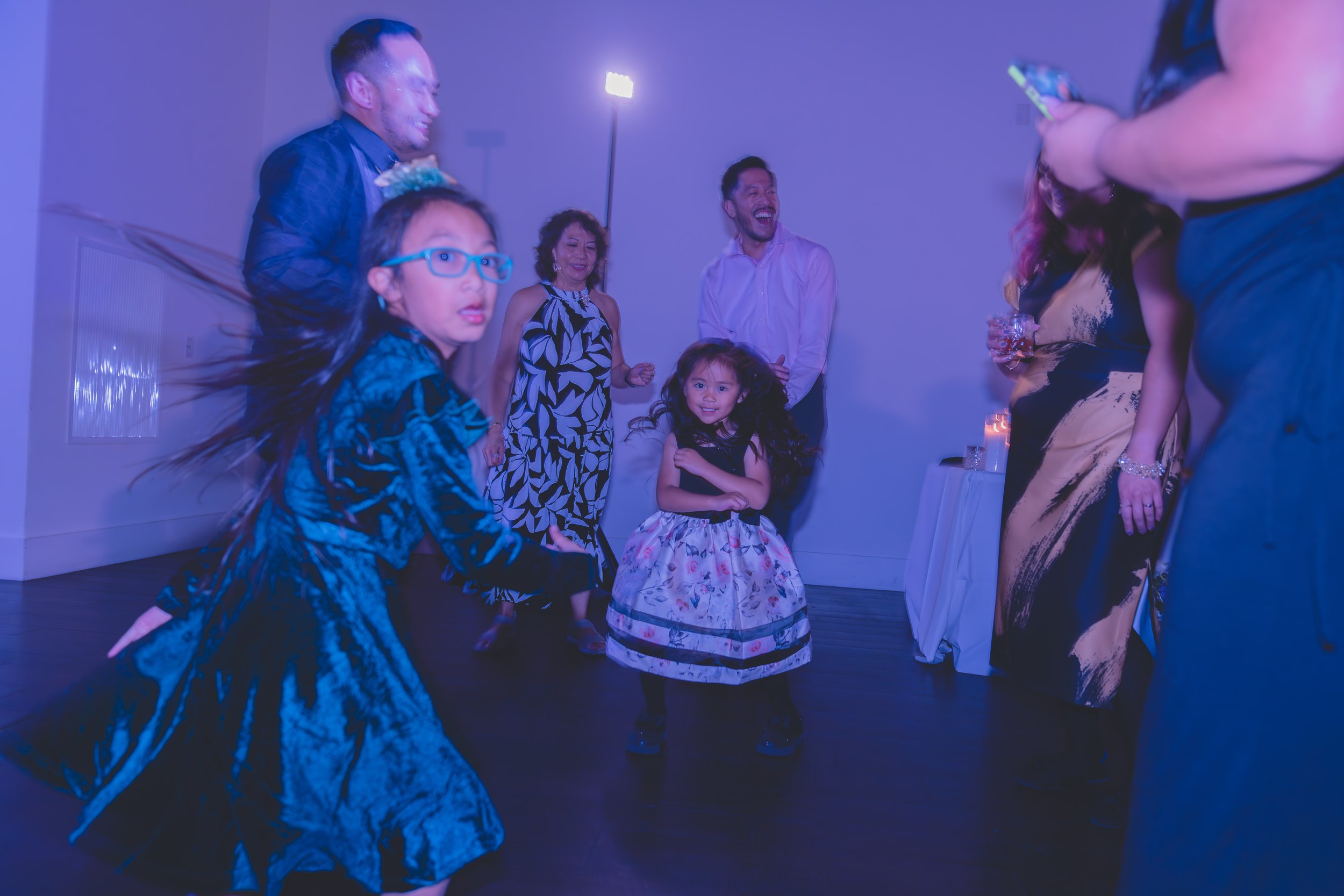 People dancing at a party, including adults and children, with blue lighting and a festive atmosphere.