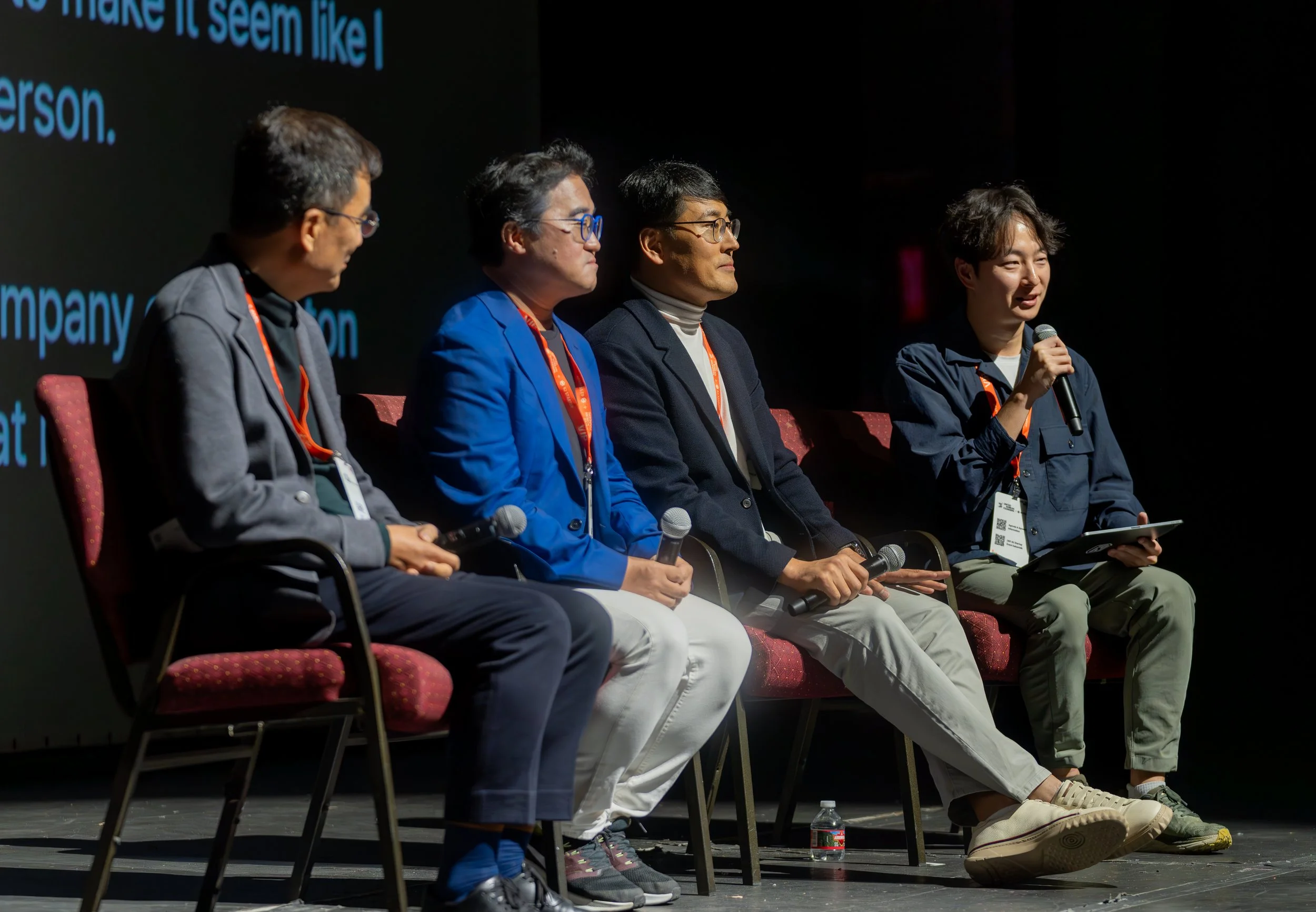 Four individuals seated on a stage during a panel discussion, each holding a microphone, with a visible screen projecting text in the background.