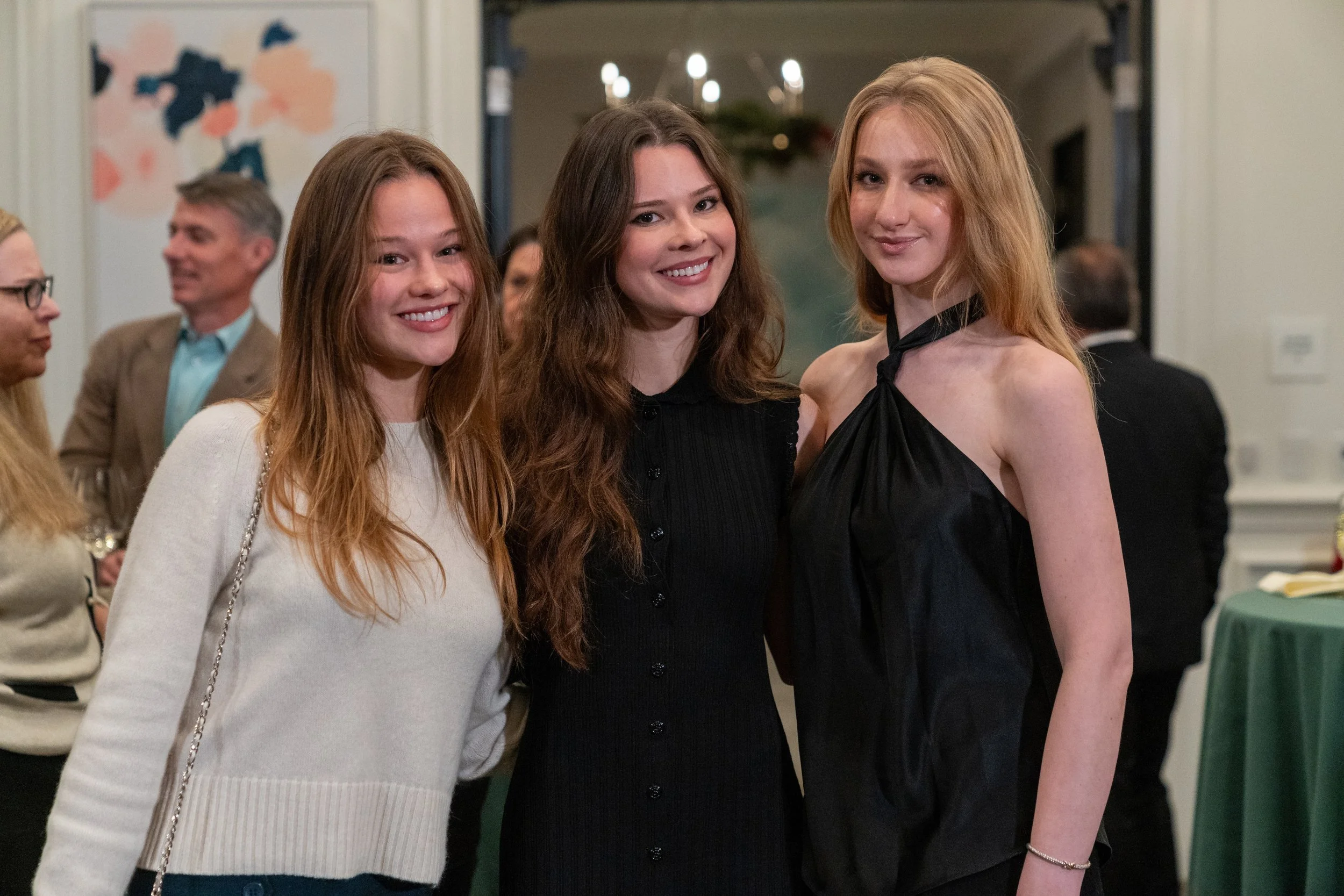Three women smiling at a social event with other people in the background.