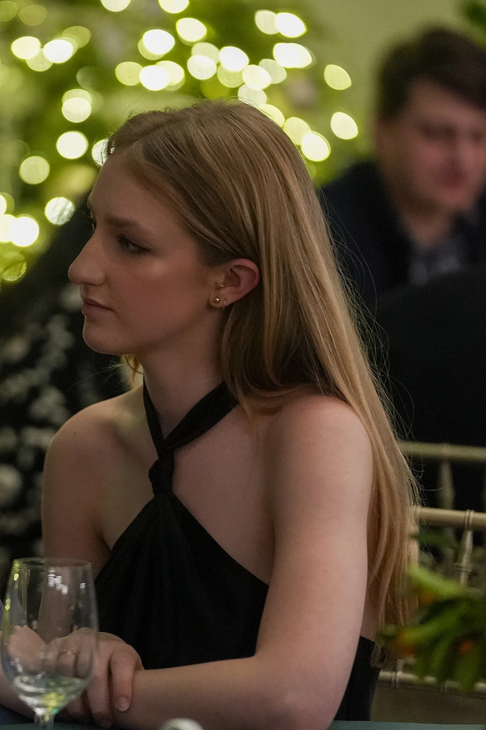 Woman in black dress sitting at table with wine glass, bokeh lights in background.