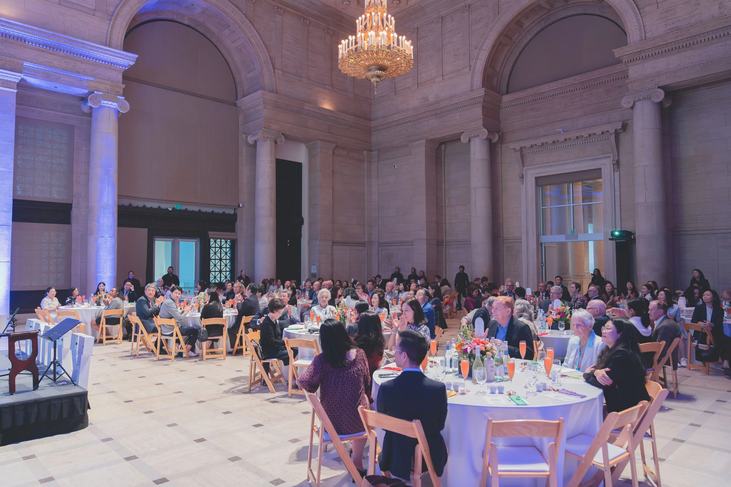 People attending a formal event or banquet in an elegant, high-ceilinged hall with large columns and a chandelier, seated at round tables decorated with flowers and tableware.