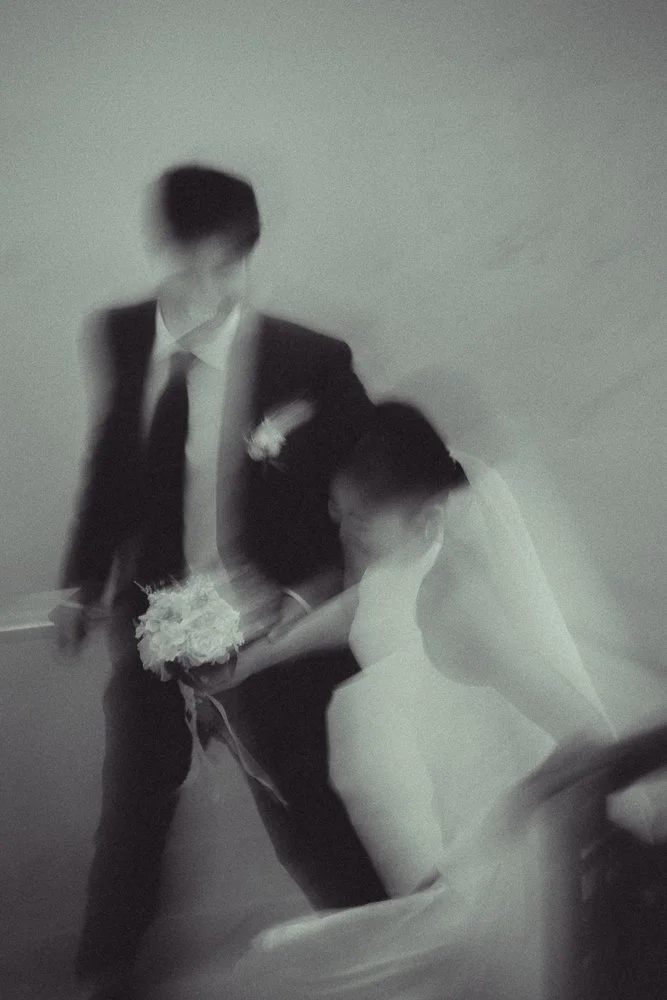 Blurred image of a bride and groom, with the bride in a white dress holding a bouquet and the groom in a suit.