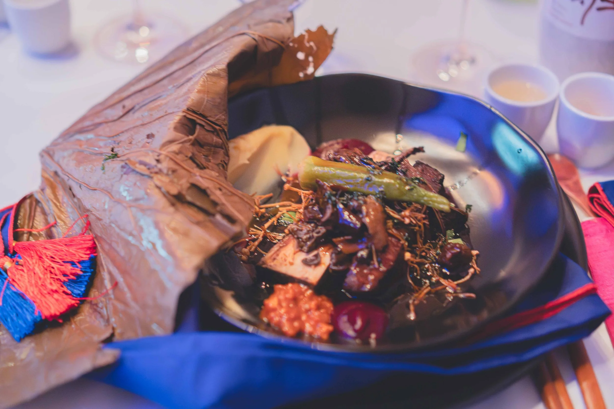 Mexican dish featuring grilled meats, vegetables, and garnishes in a black bowl, with tortilla wrapped in paper on the side, on a table with glasses and small bowls in the background.