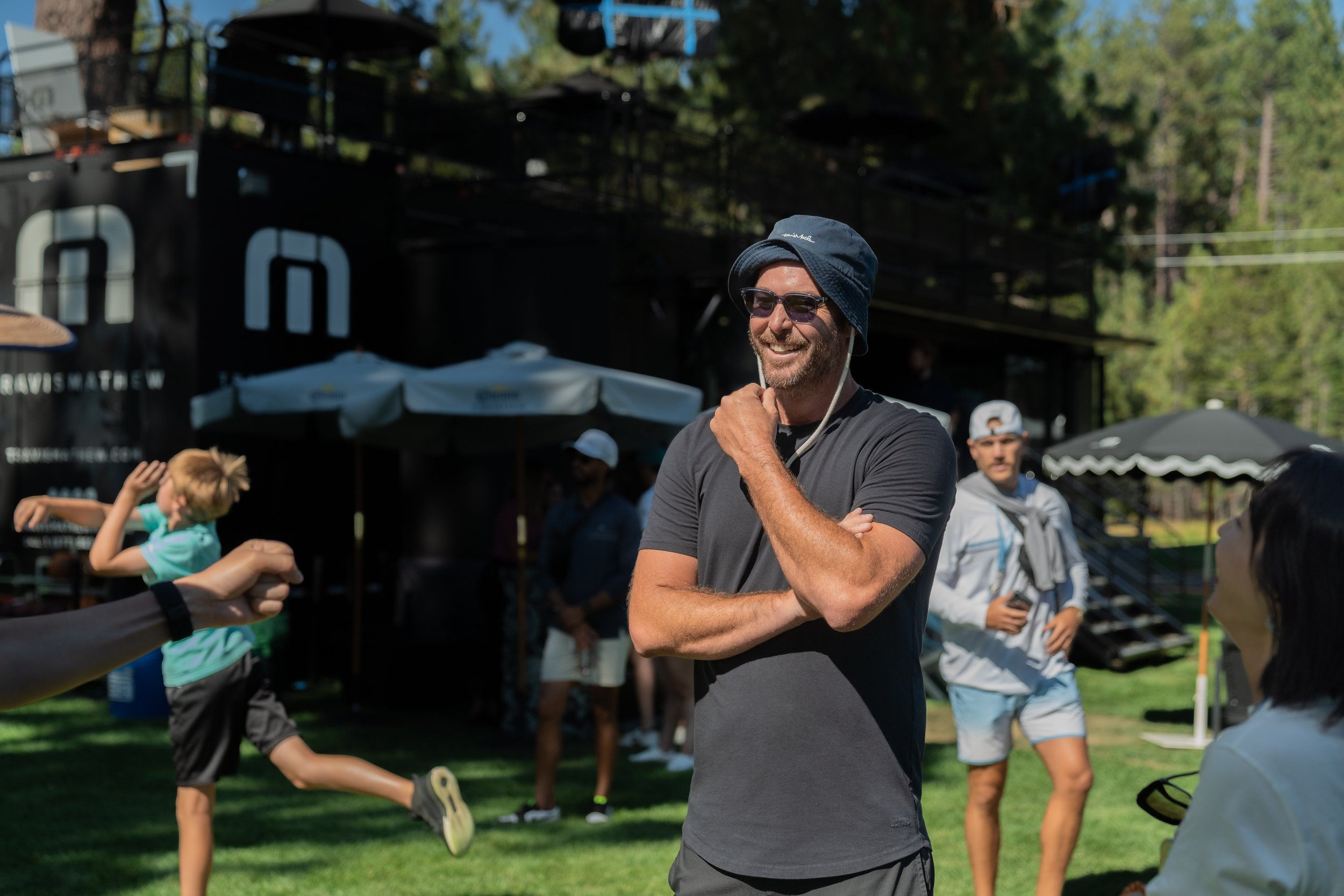 A group of people outdoors in a sunny setting. The focus is on a smiling man wearing a bucket hat and sunglasses, with his arms crossed. Others are casually dressed and participating in various activities. There are umbrellas and a backdrop with the