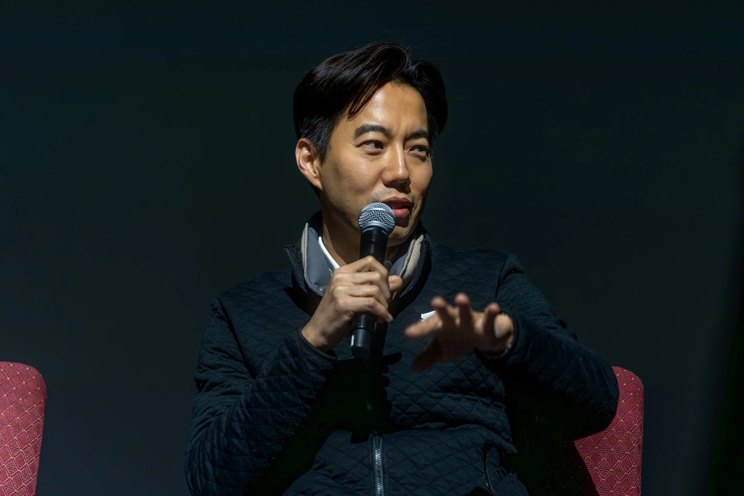 Person speaking into a microphone while seated on a chair.