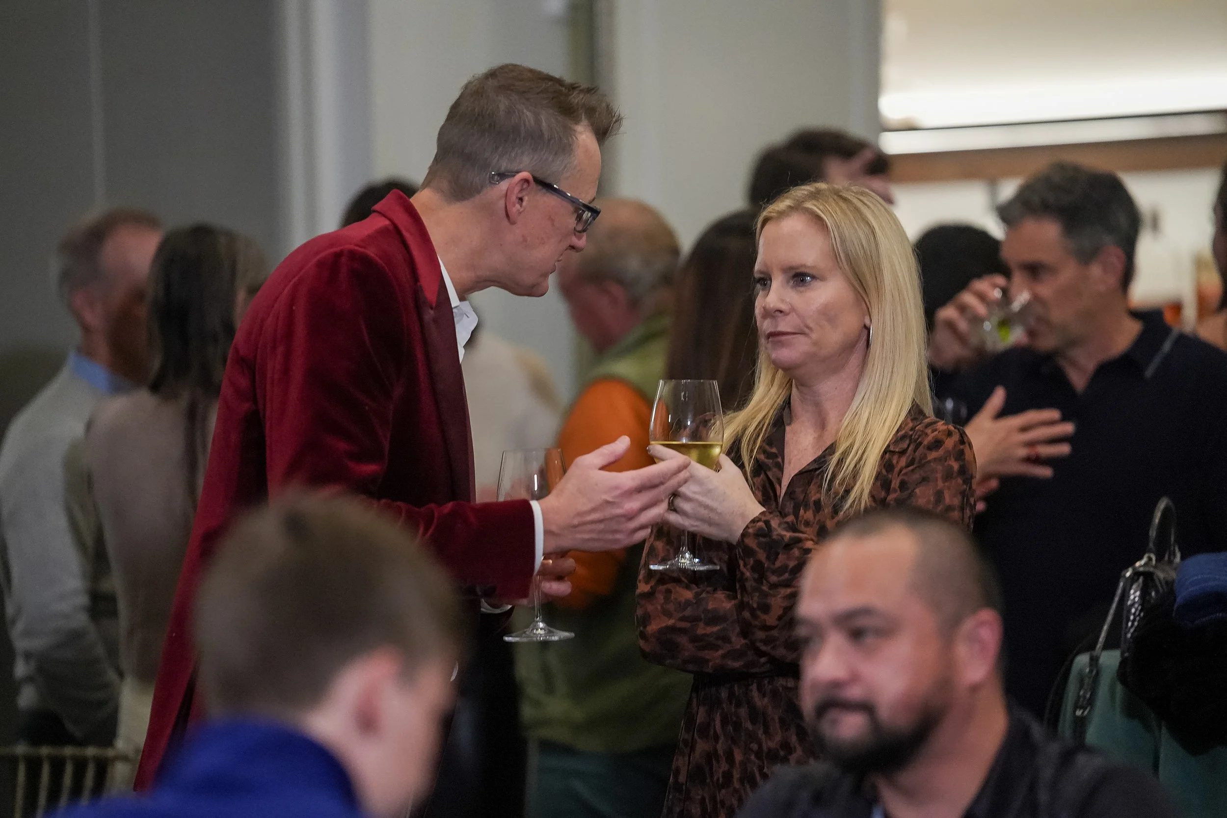 People socializing at a gathering, holding wine glasses, with a man in a red jacket talking to a woman in a patterned dress. Other attendees are seen in the background.