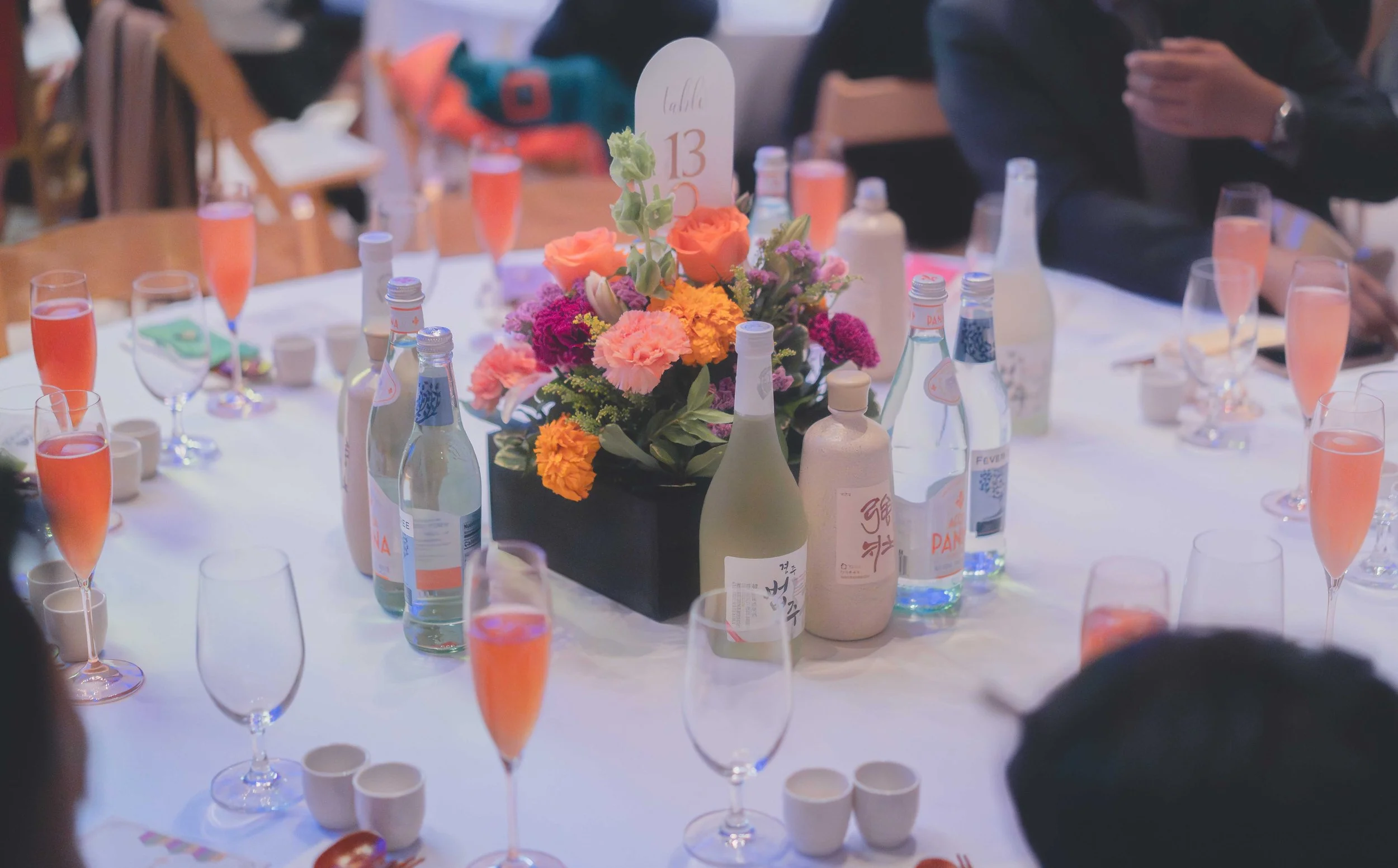 Table set for a celebration with a centerpiece of pink, orange, and purple flowers, bottles of sparkling water, glasses of pink champagne, and cups, with people in the background.