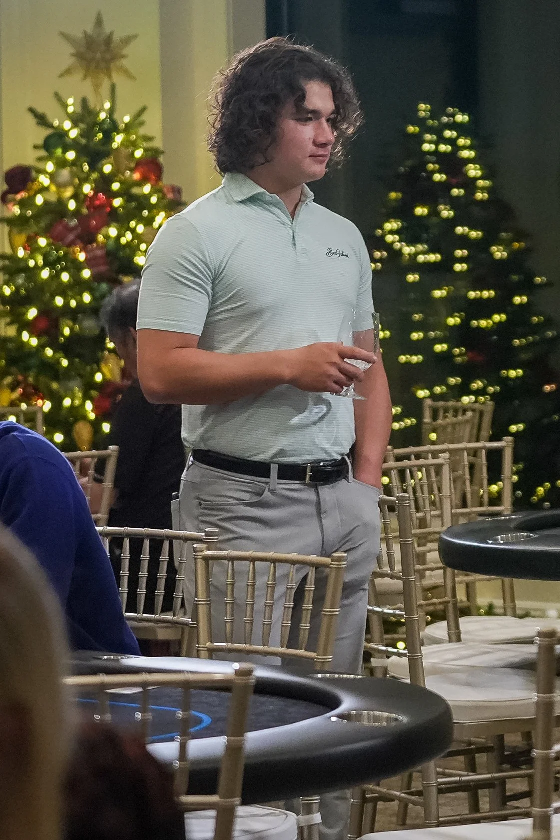 Man in light polo shirt holding a glass, standing near decorated Christmas trees, with chairs and a table in the foreground.
