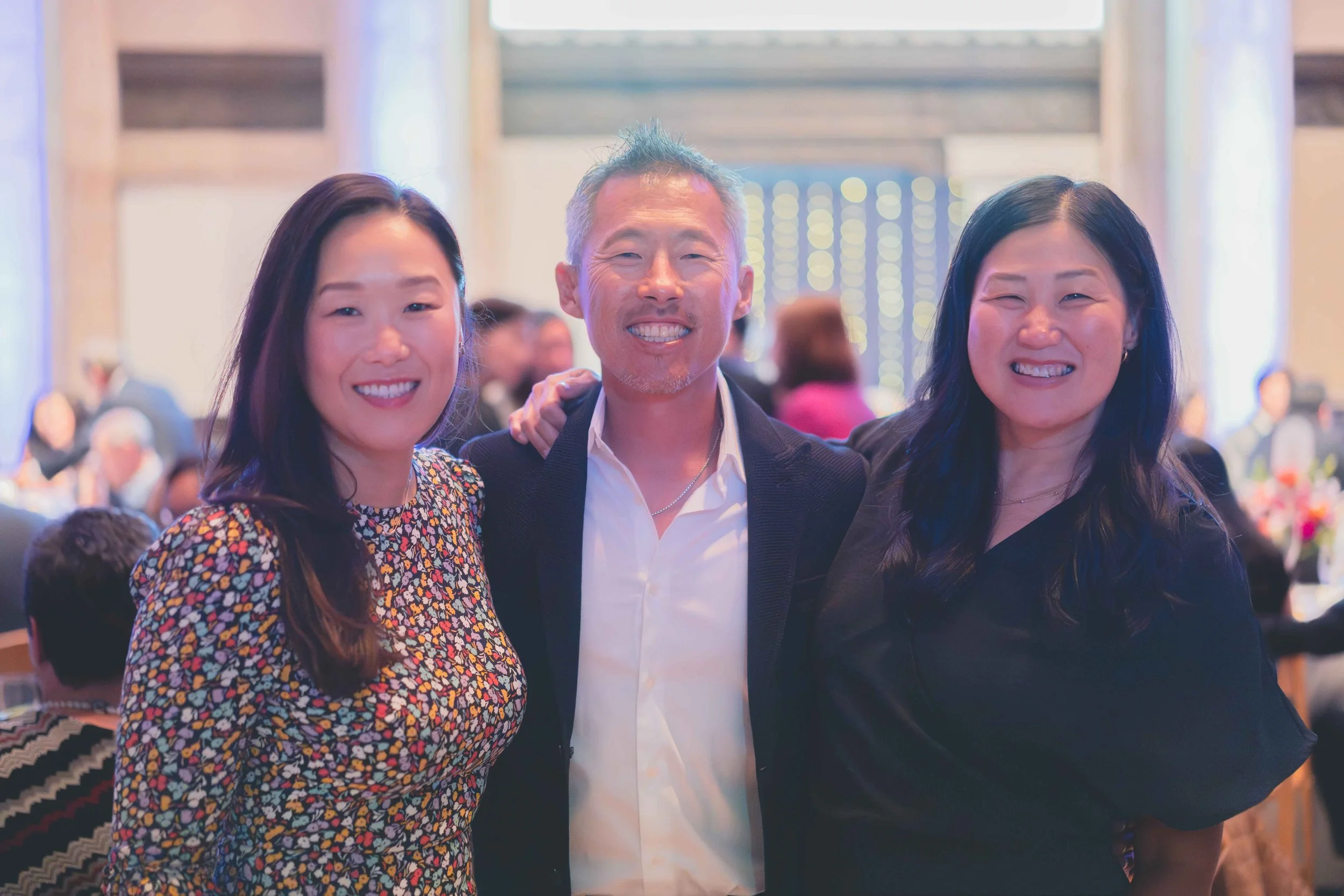 Three people smiling at a social event, standing close together, with a blurred background of people and decorations.