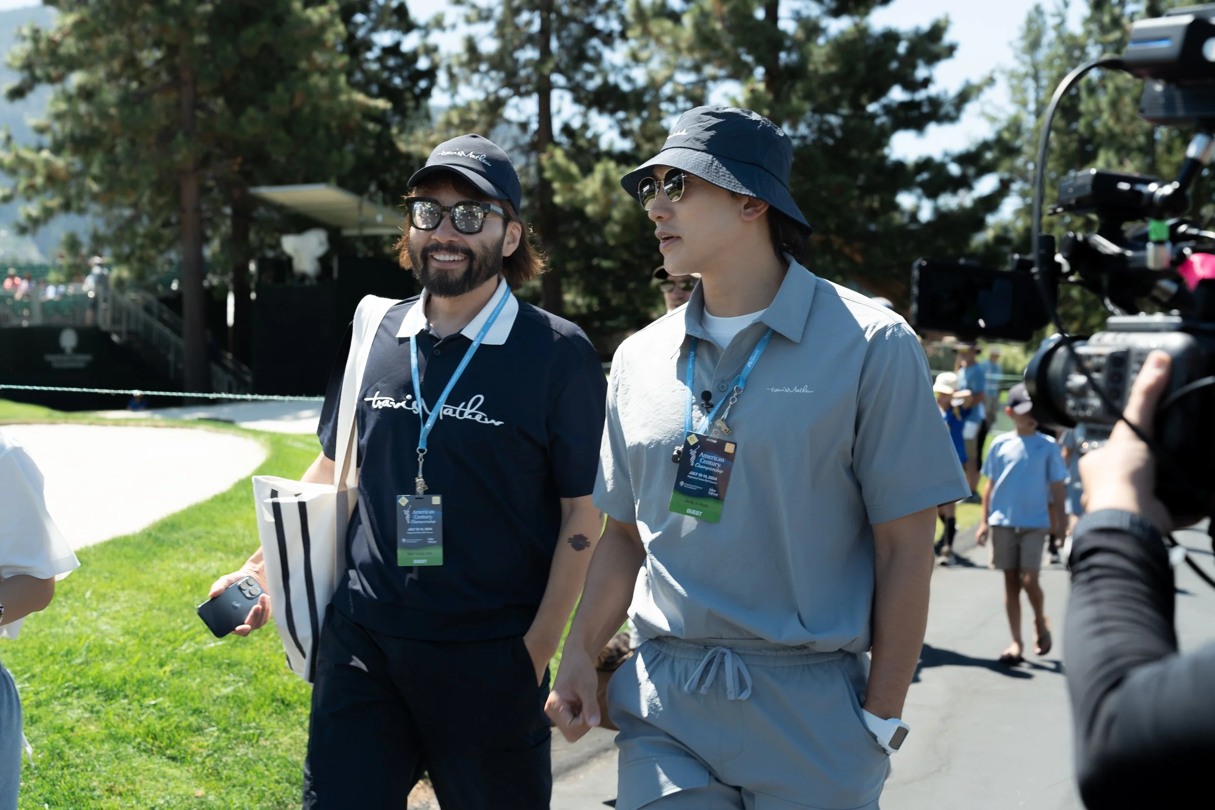 Two people walking outdoors at a golf event, wearing sunglasses and casual sporting outfits with lanyards. A camera is filming them, with trees and greenery visible in the background.