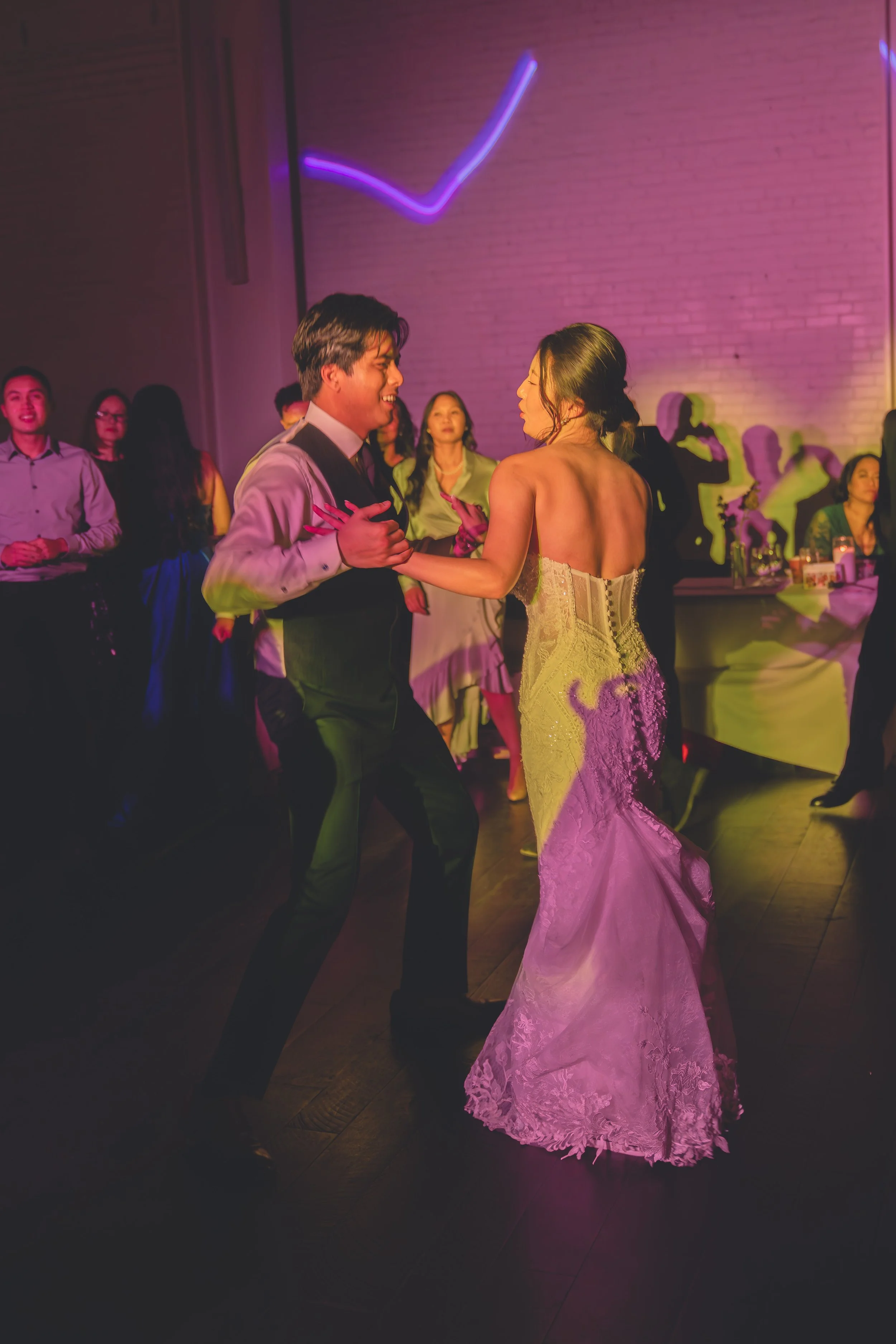 A couple dancing at a wedding reception with guests watching, in a dimly lit room with colorful lights.
