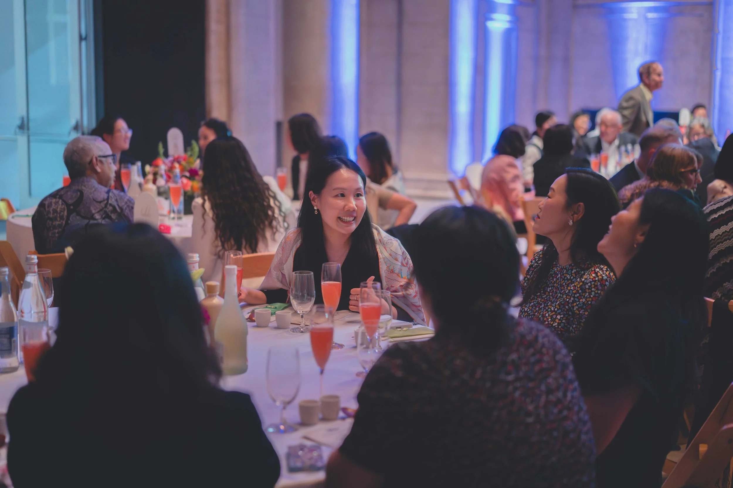 People sitting at round banquet tables in elegant attire at a formal event, engaging in conversation, with a woman smiling and talking to others, and a woman in patterned dress listening, set in a grand hall with blue lighting and stone columns.