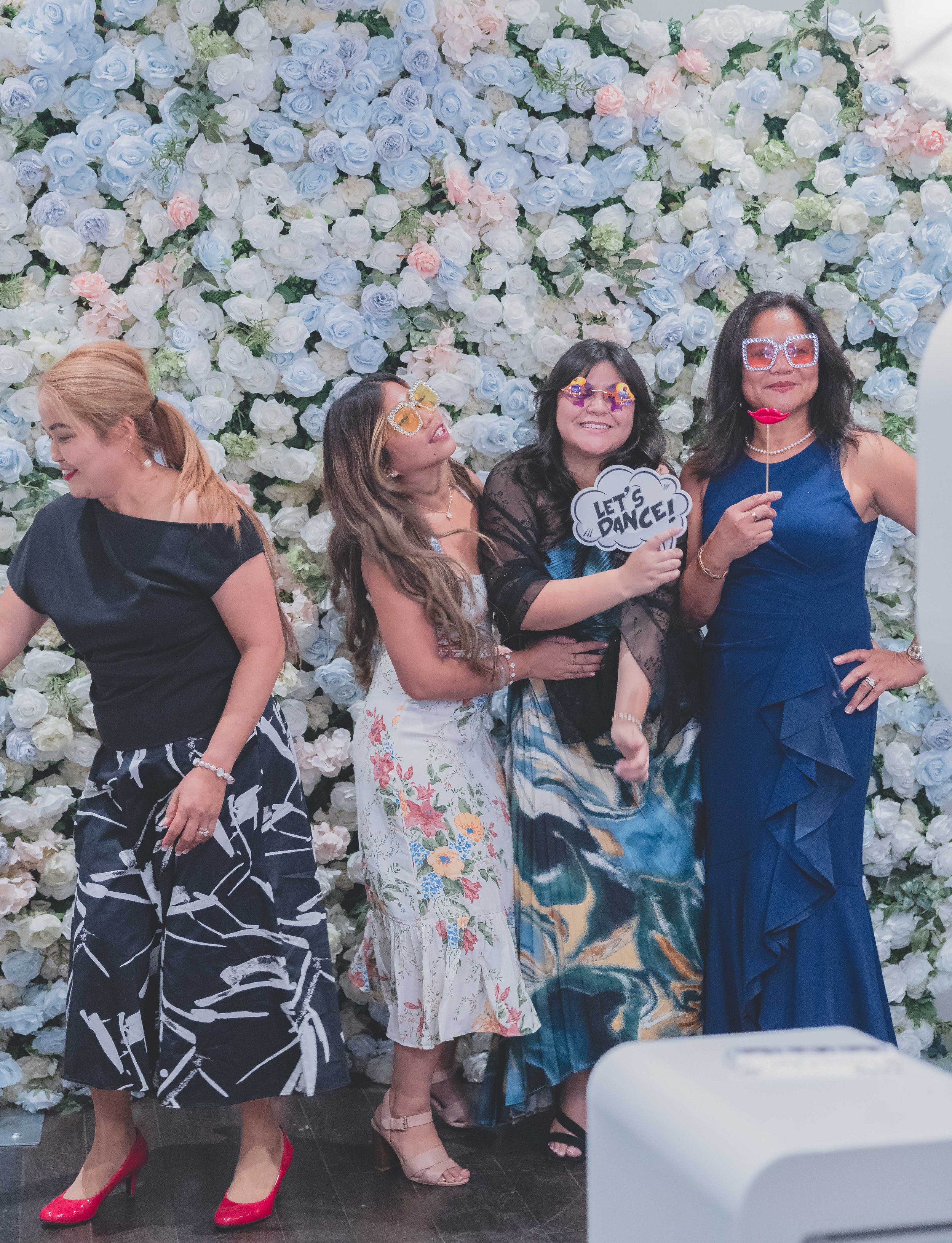 A group of four women posing in front of a floral backdrop, featuring white and pastel-colored roses. They are wearing stylish dresses and sunglasses, holding playful props like a "Let's Dance!" sign. The mood is festive and cheerful.