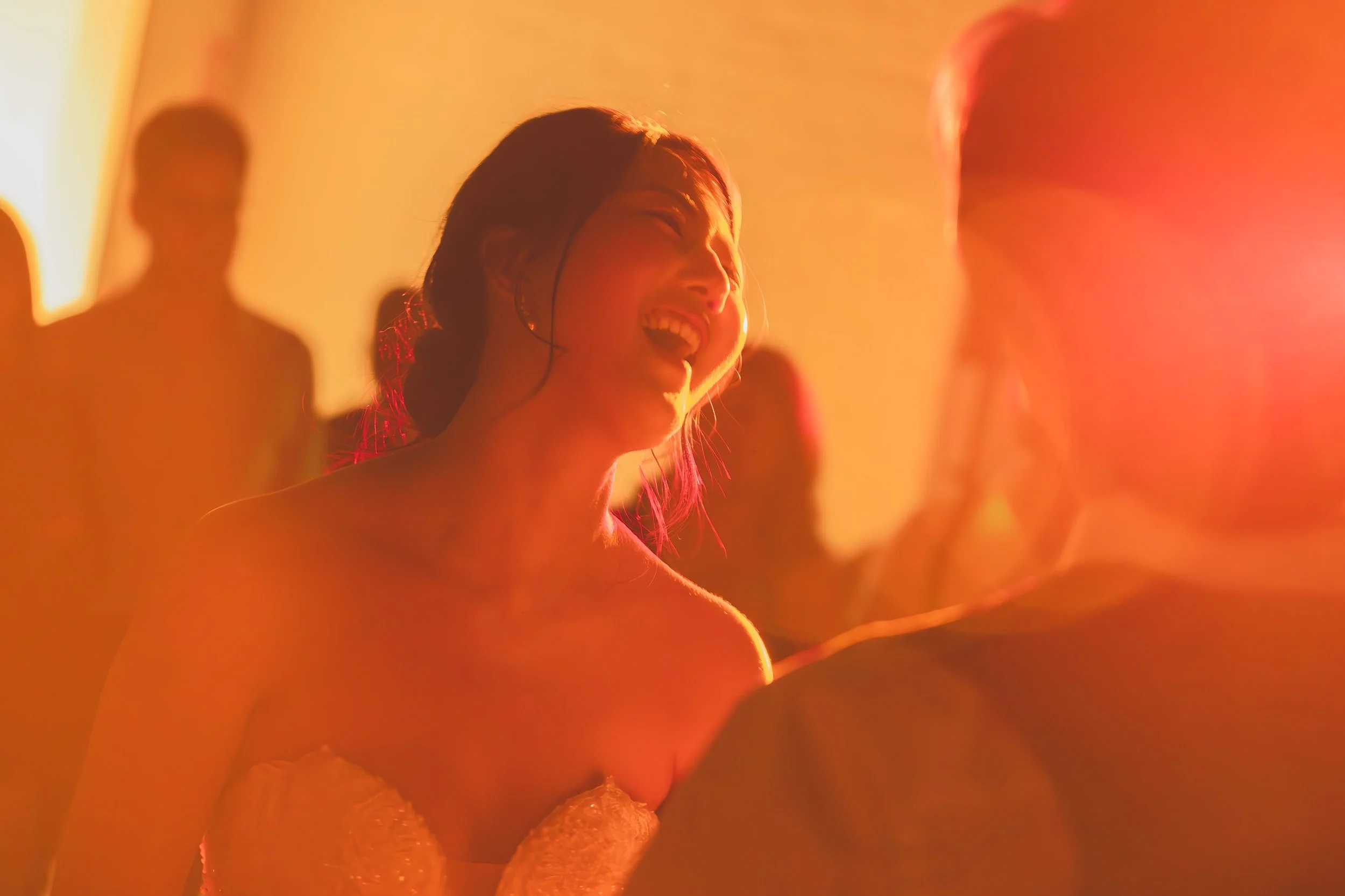 Woman laughing indoors with a warm, golden glow, surrounded by blurred people.