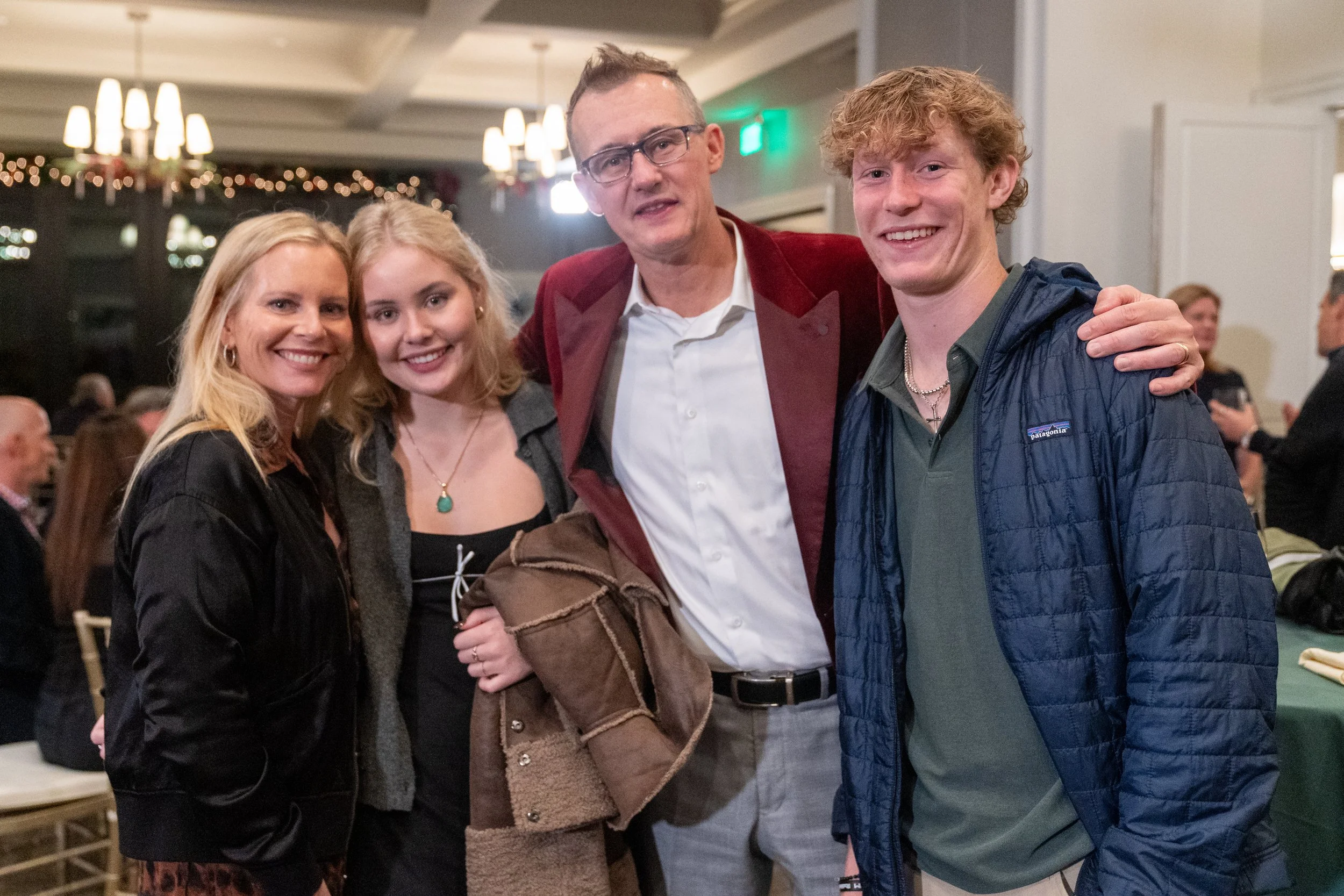 Group of four people smiling and posing together at an indoor event, with decorative chandeliers and holiday lights in the background.