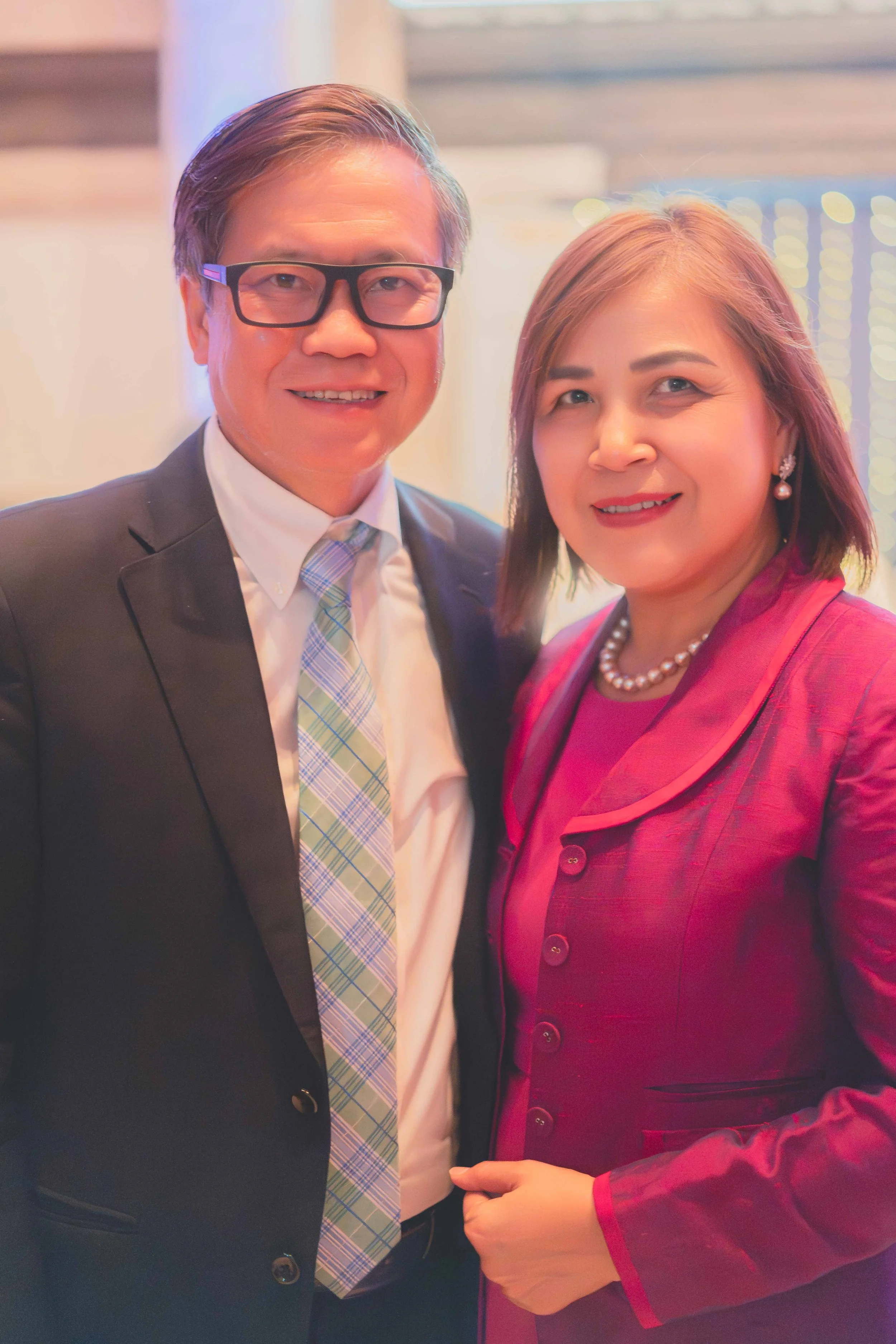 A man and woman dressed in formal attire smiling at the camera, indoors with blurred background lights.
