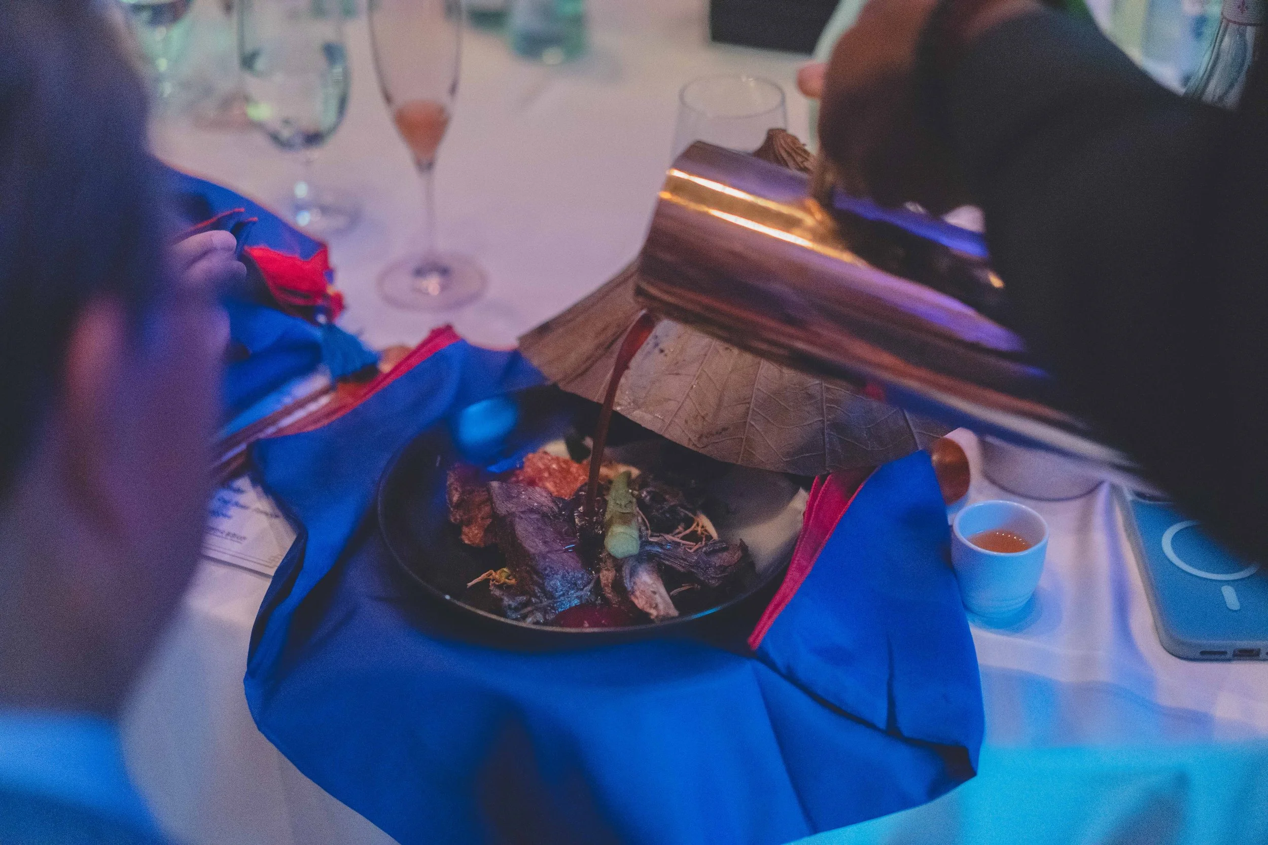 A person pouring gravy onto a plate of steak and vegetables at a dining table in a dimly lit setting with glasses of drinks in the background.